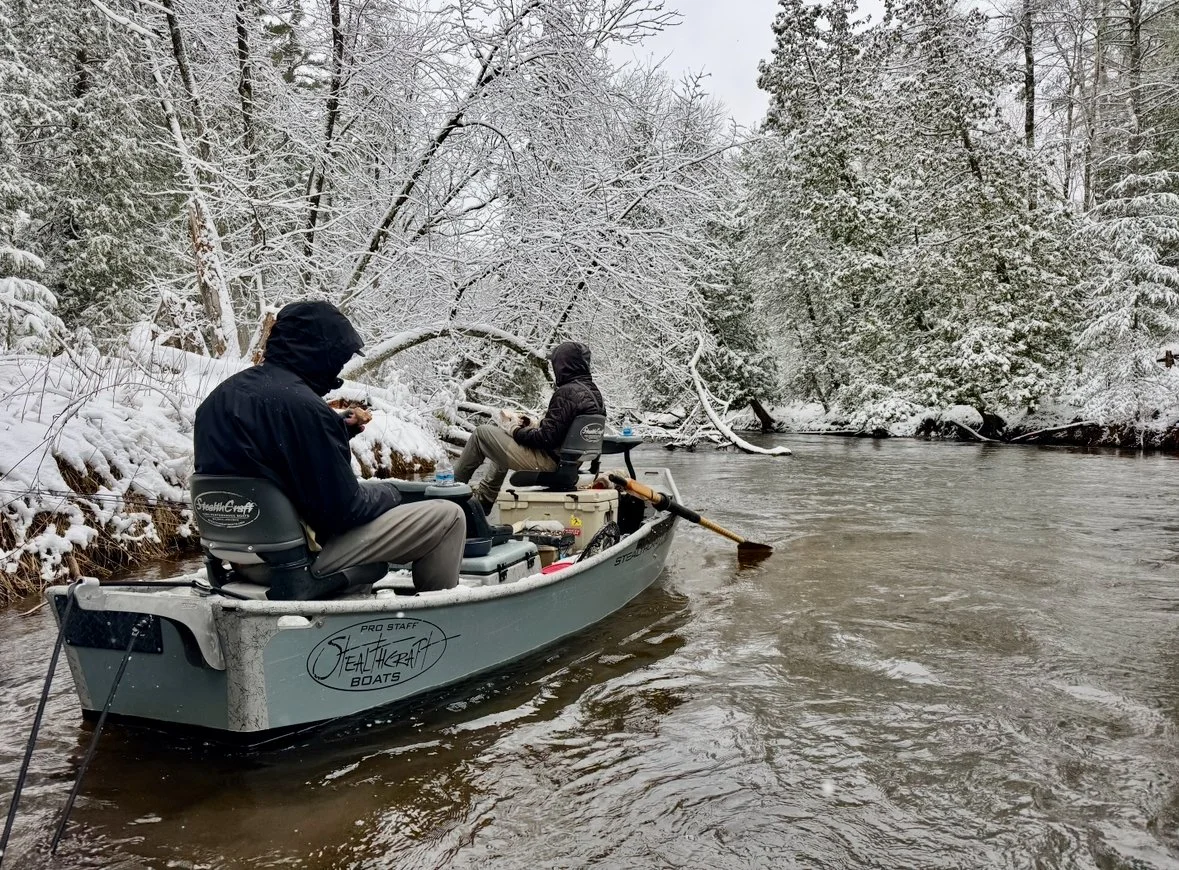Two people in a small boat on a river with snow-covered trees along the banks, wearing dark jackets and hoods, with one person using a paddle.