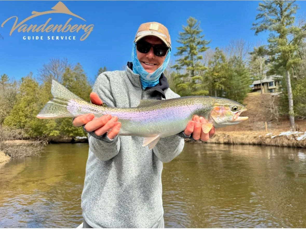 Person holding a rainbow trout fish by a river, with trees and a house in the background, under a blue sky.