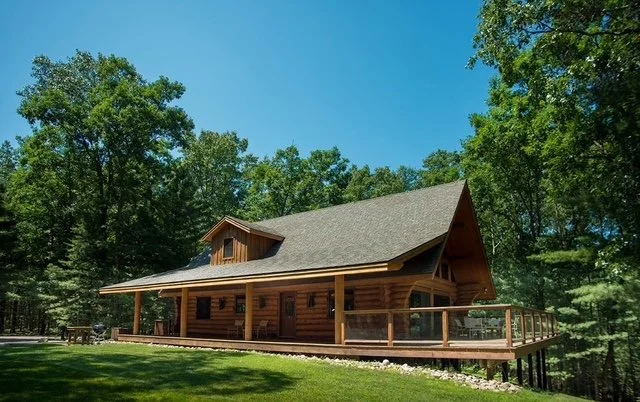 A wooden house with a steep roof, large porch, surrounded by green trees and a well-maintained lawn under a clear blue sky.