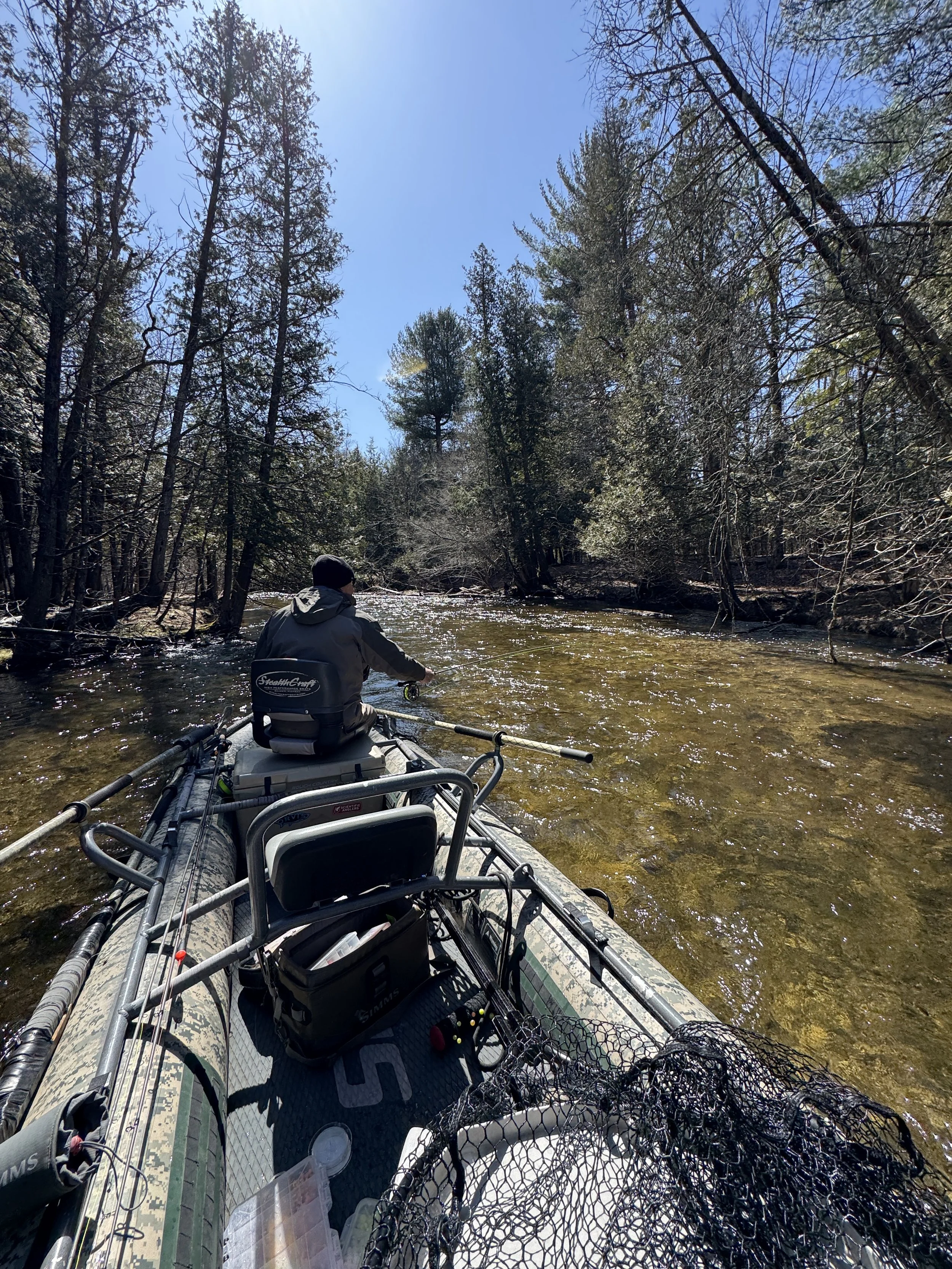 Person fishing from a boat on a river surrounded by trees on a clear, sunny day.