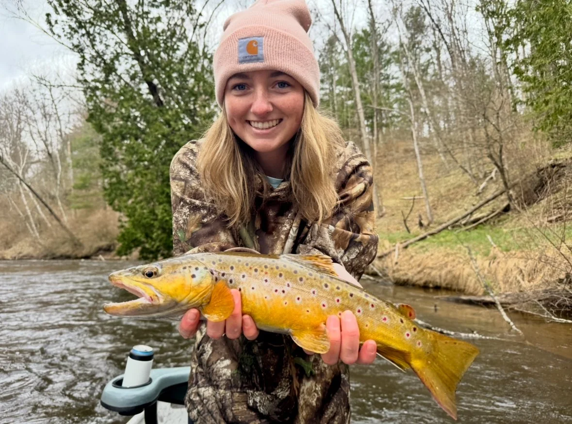 A young woman in camouflage jacket and pink Carhartt beanie smiling and holding a large rainbow trout by the riverbank.