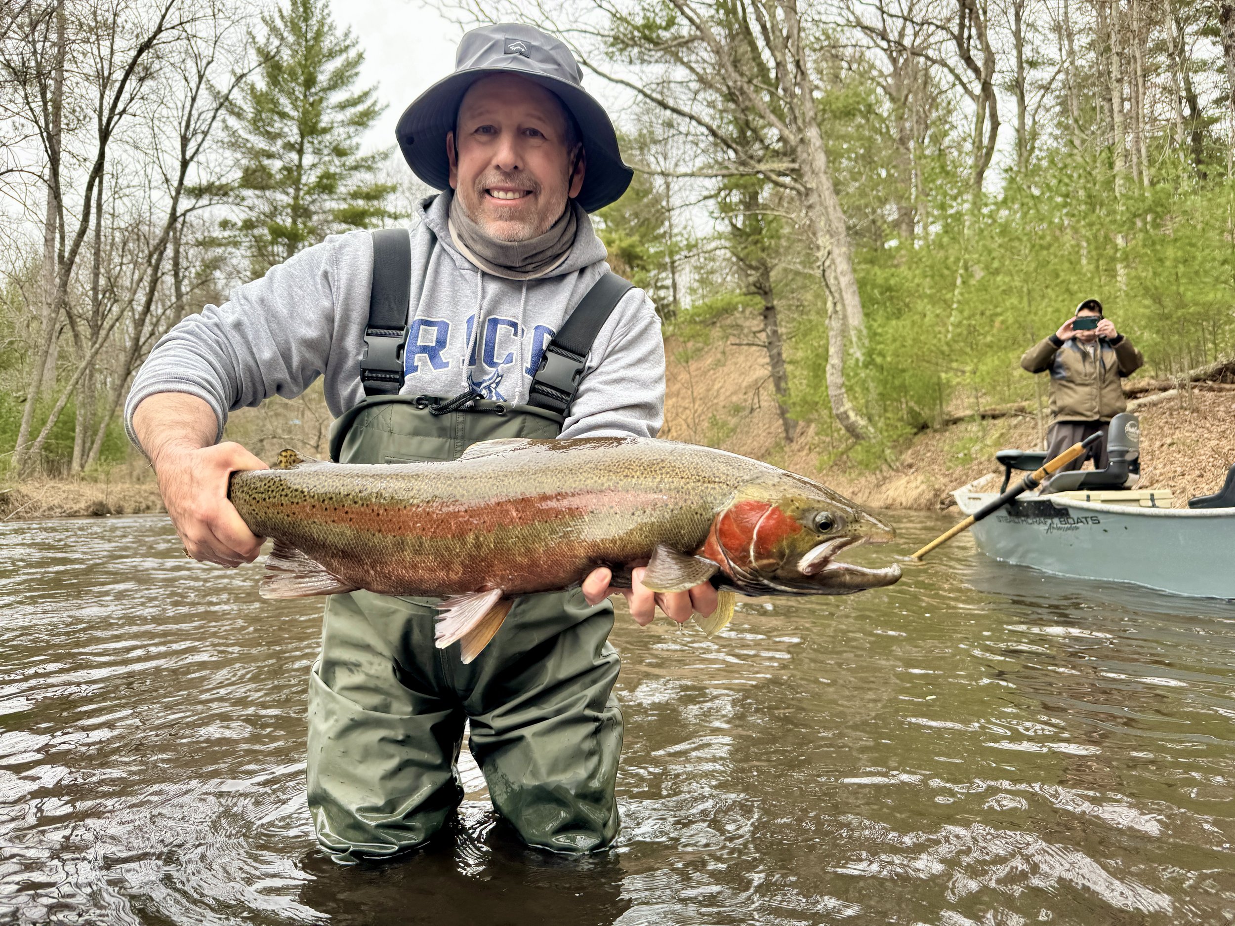 A man wearing a gray hoodie and camouflage waders holding a large rainbow trout in a river, with another person taking a photo on a boat in the background.
