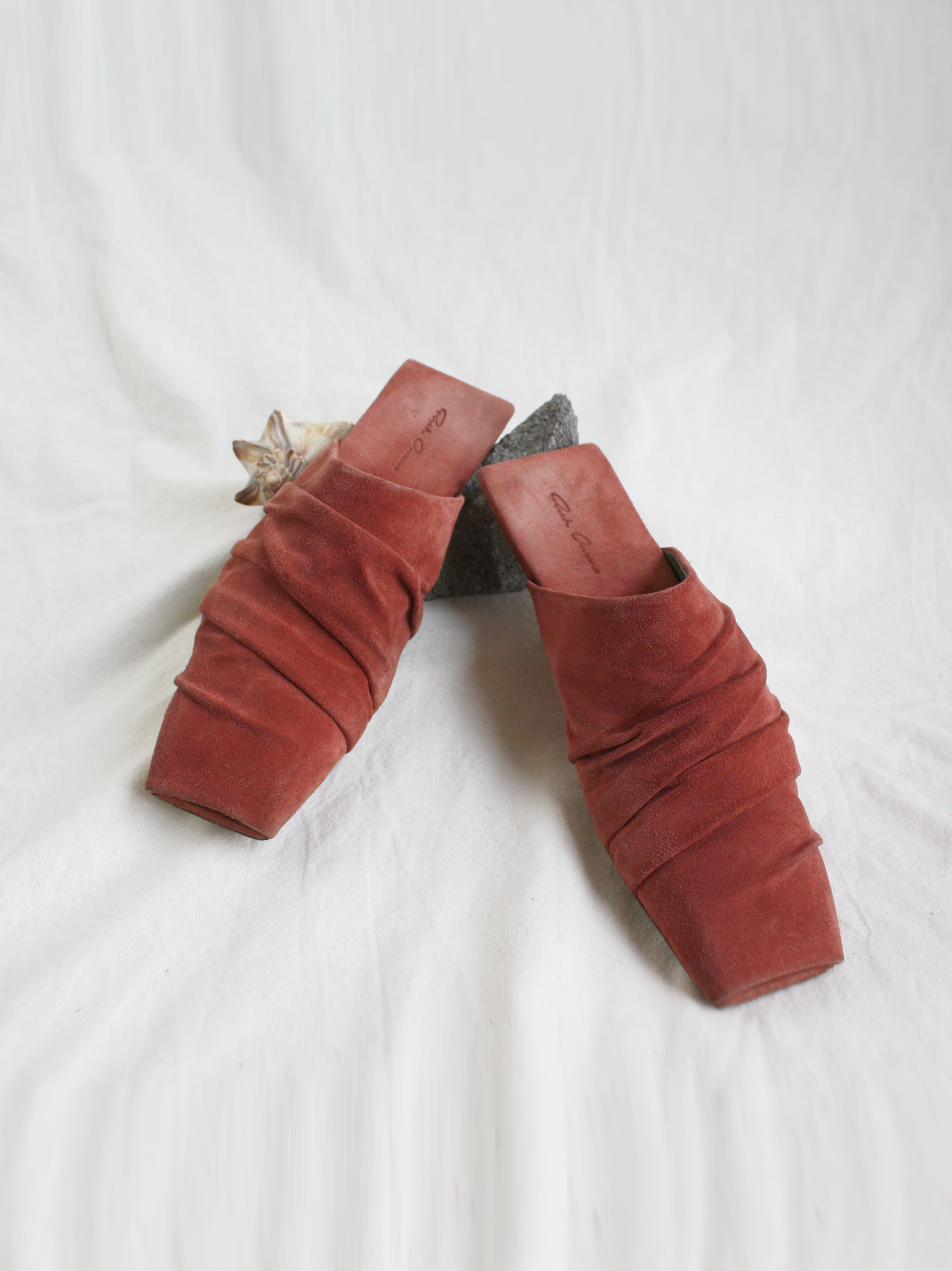 Burnt Pink Draped Leather Slippers with square toe and heel on a white muslin backdrop with a shell and concrete