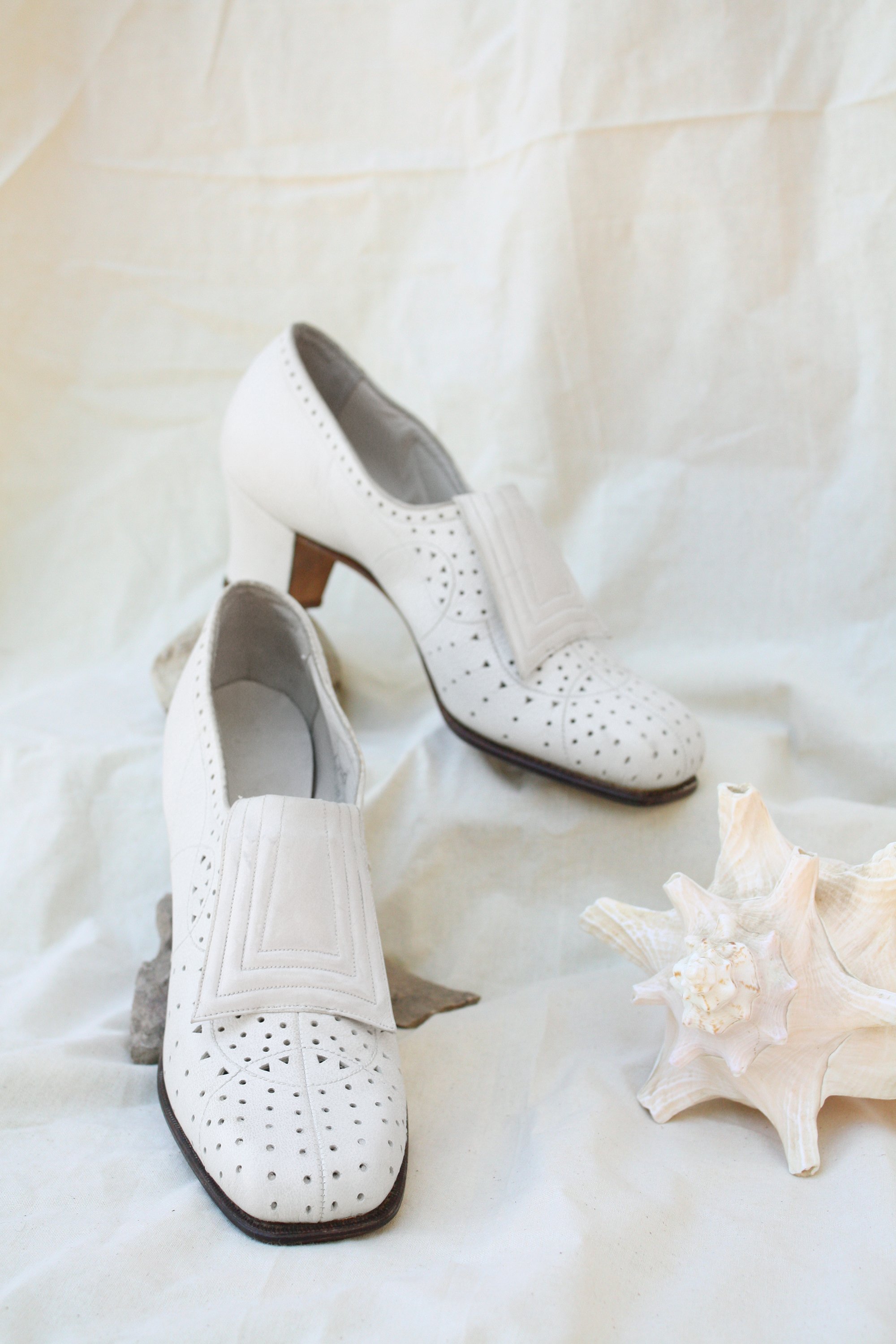 1960s white pinhole leather loafers with block heel and stylized tongue against a muslin backdrop with seashell