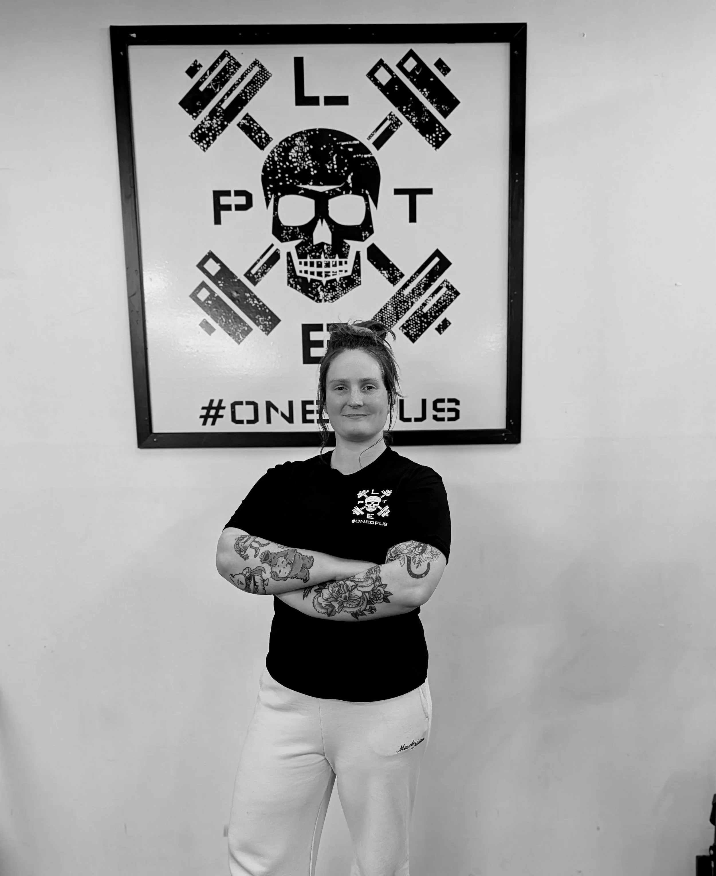 Black and white photo of a woman with tattoos on her arms, wearing a black shirt with a skull logo and white pants, standing in front of a wall with a graphic design featuring a skull, crossbones, and the words 'LIFT POST' and '#ONEOFUS'.