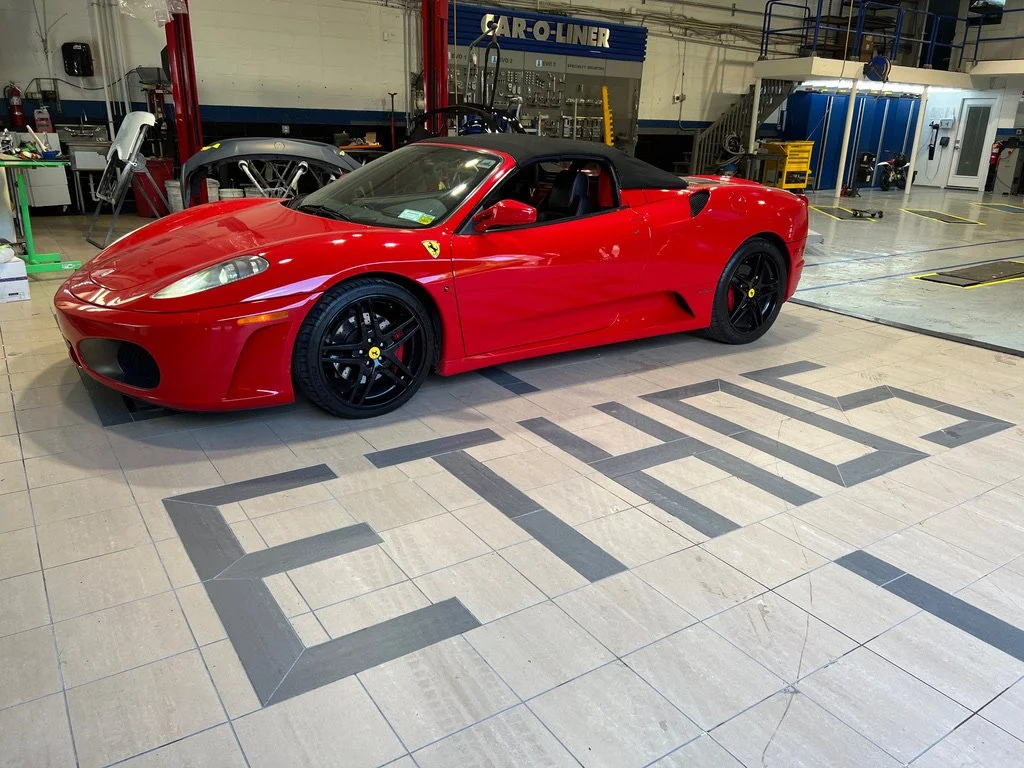 A red Ferrari sports car with black wheels inside a workshop or garage, with 'Ferrari' written on the floor in front of it.