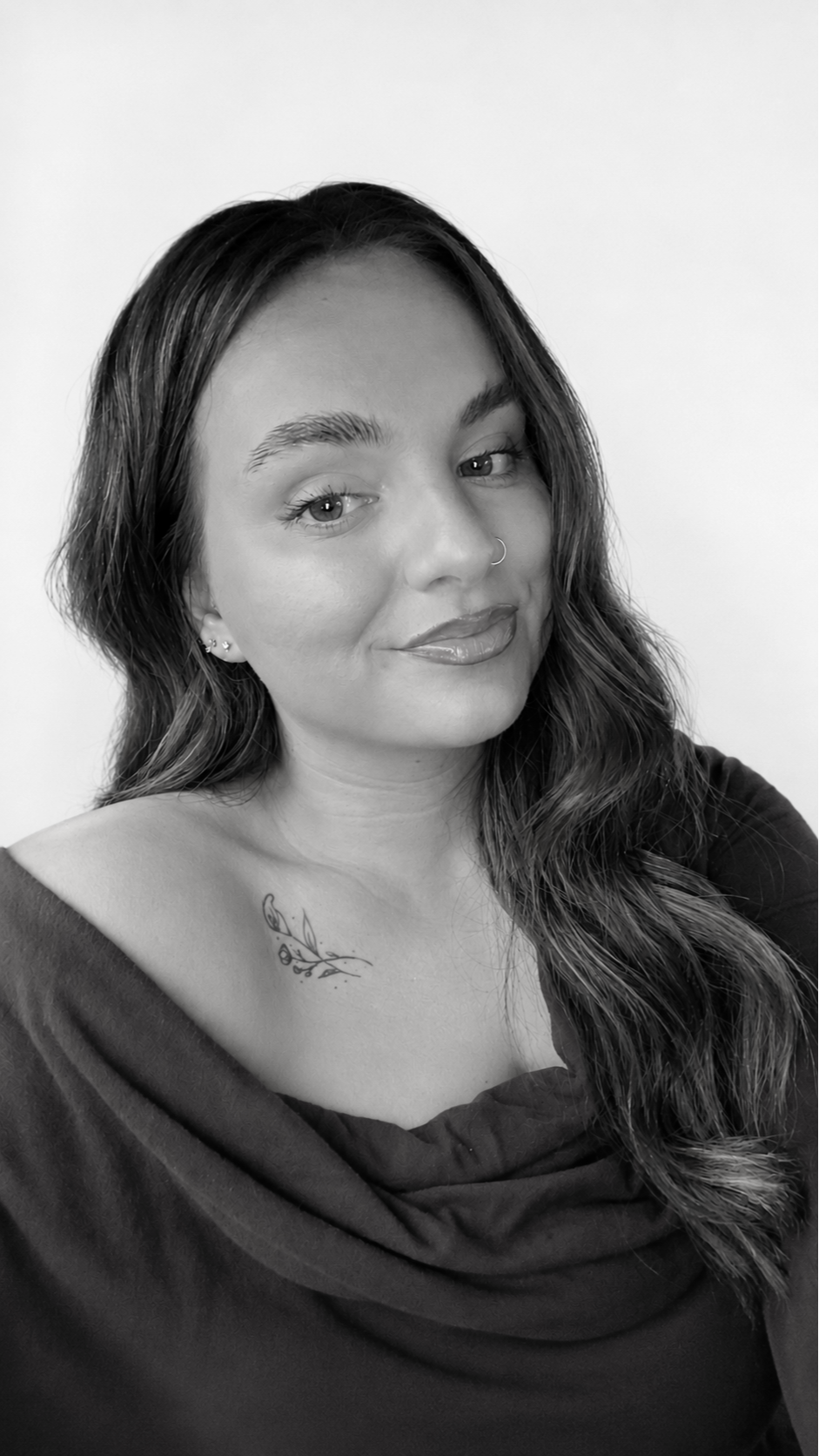 A black-and-white portrait of a woman with wavy hair, a nose ring, and a tattoo on her shoulder. She is wearing a dark top and has a slight smile, looking at the camera.