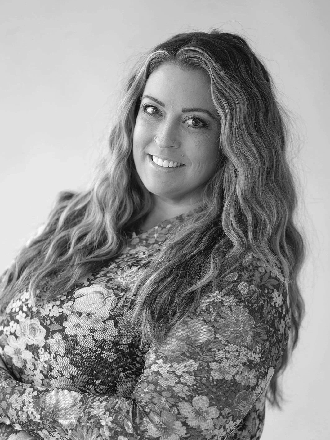 Black and white photo of a woman with long wavy hair smiling at the camera, sitting indoors.