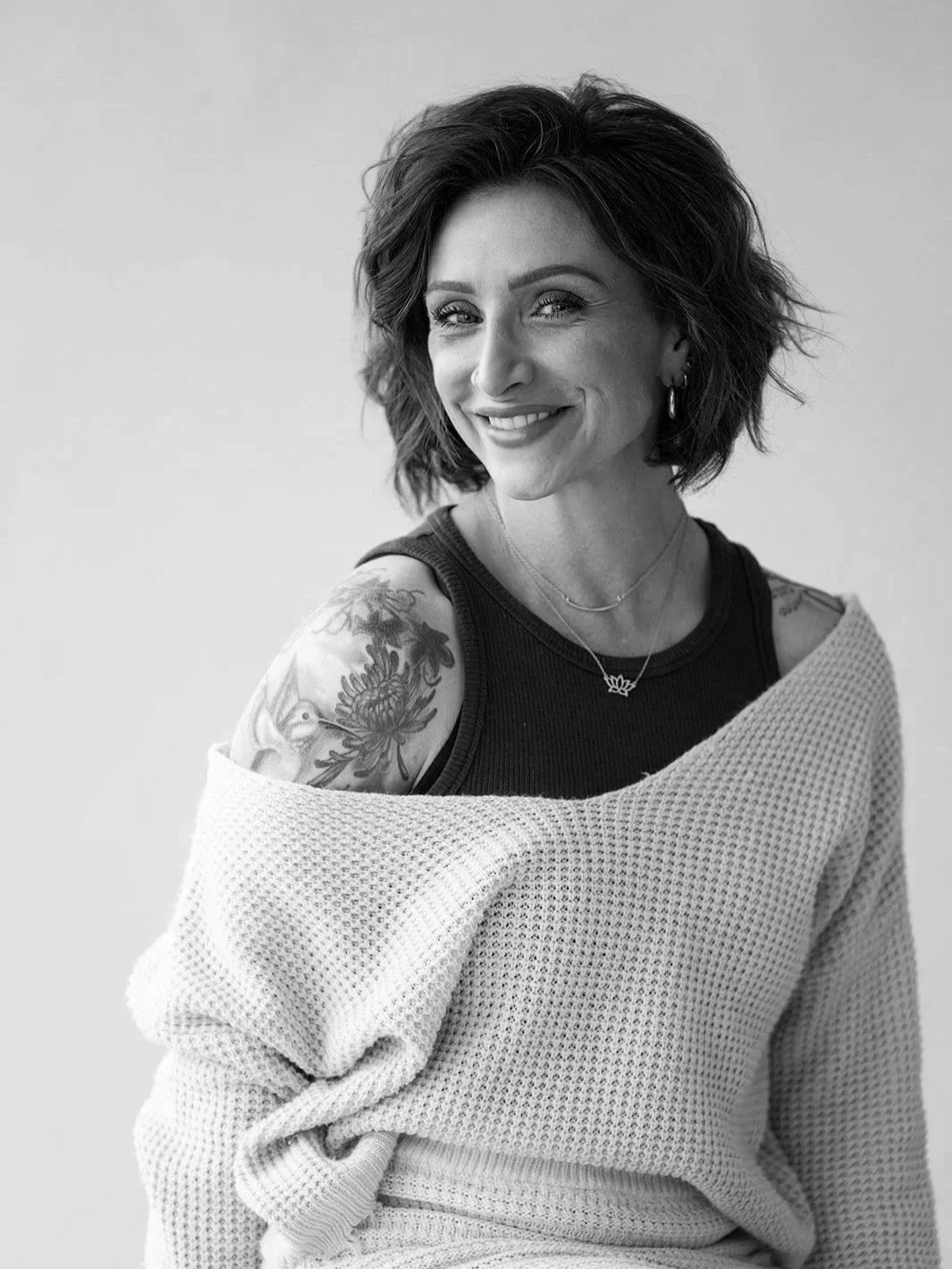 A woman with shoulder-length dark hair, wearing a black top and hoop earrings, smiling at the camera against a textured wall background.