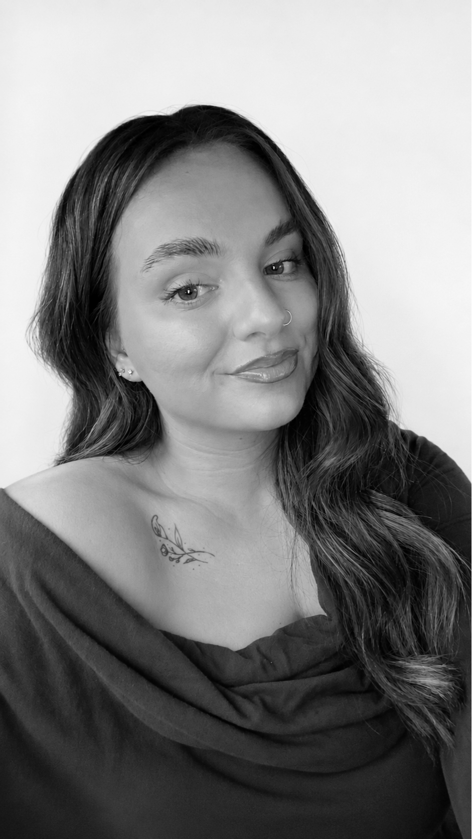 A woman with long, wavy hair and a nose ring, wearing a dark top and a tattoo on her left shoulder, posing against a plain background.