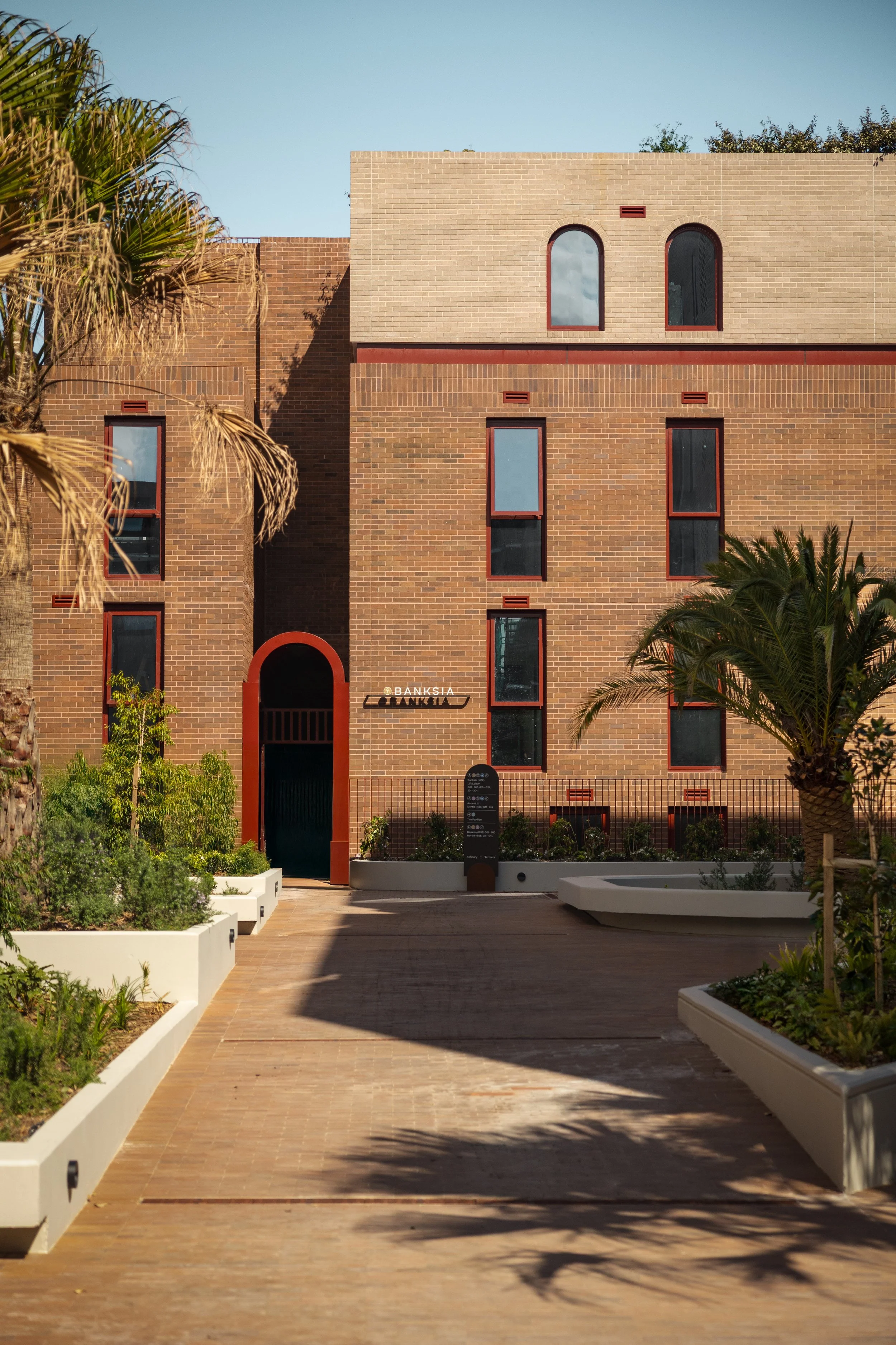 The image shows a modern brick building with several arched and rectangular windows, a landscaped walkway with planters, and palm trees in the foreground, under a clear blue sky.  Luxury architecture design, high end residential development