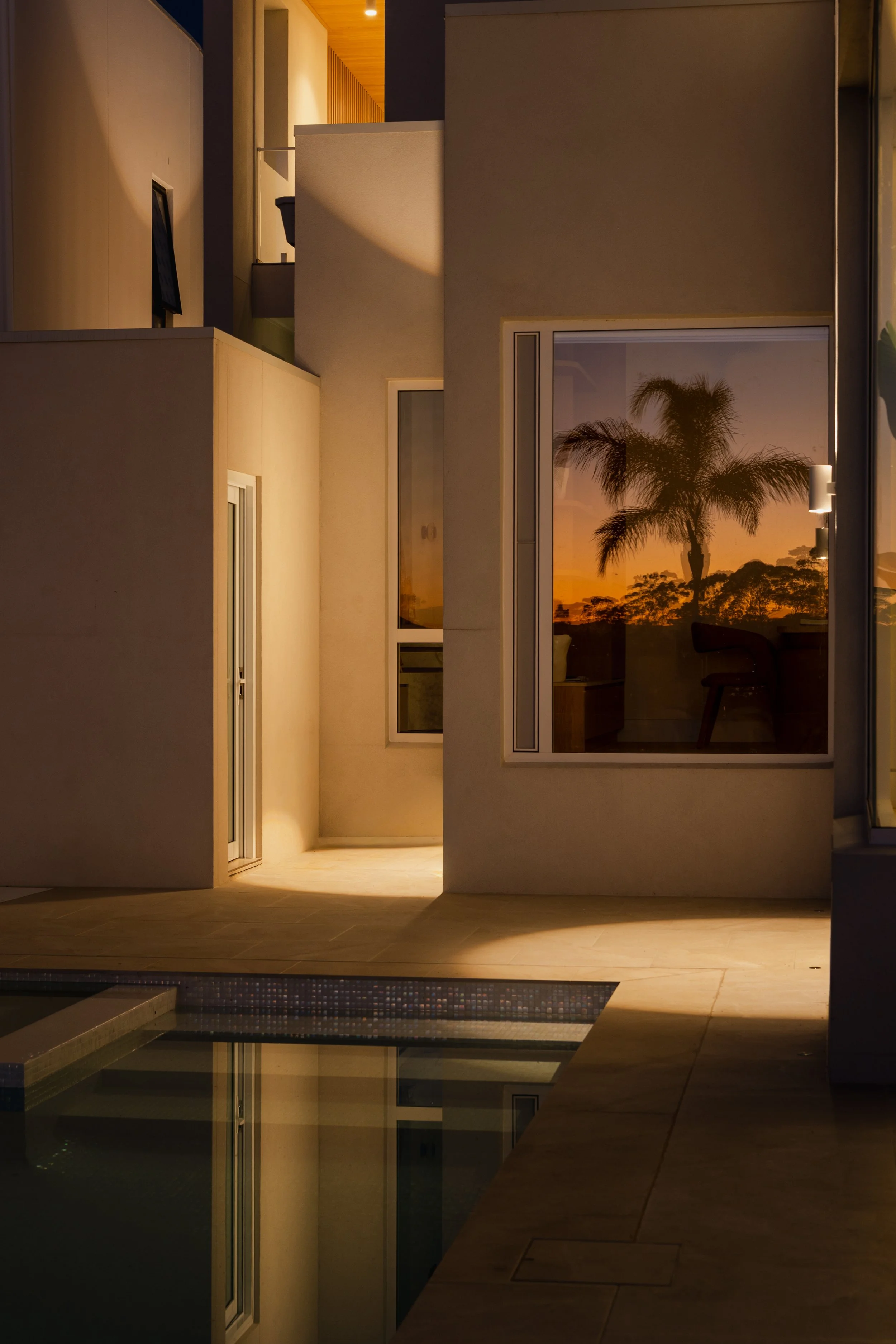 The photo shows an indoor scene at dusk or dawn, with a large window reflecting a palm tree and a sunset sky. The lit interior has white and beige walls, with visible furniture and a door, and the reflection of the sky and palm tree can be seen in th