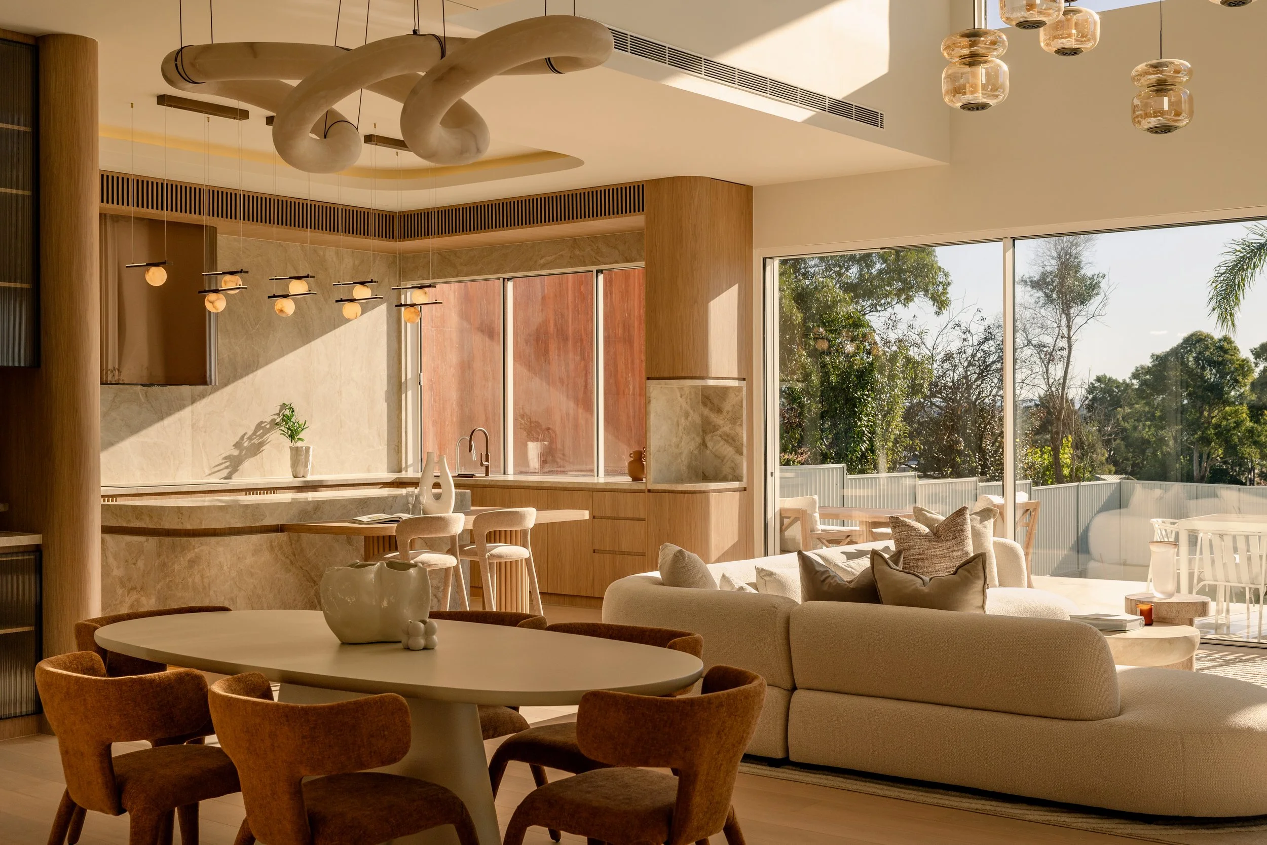 Modern living space with large windows, hanging pendant lights, a dining table with chairs, a sofa, and outdoor seating visible through the windows, with natural sunlight flooding the room.