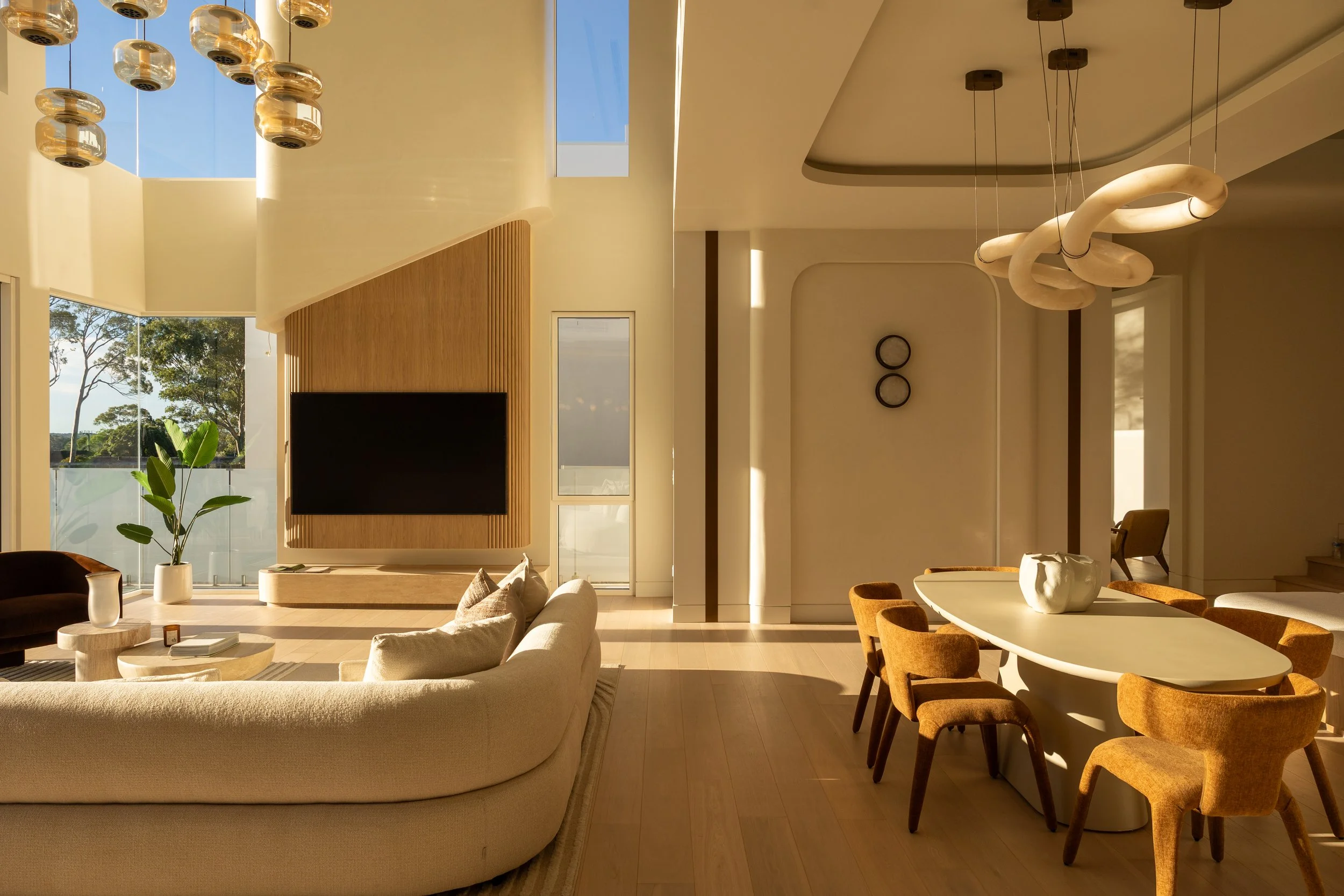 A modern living room with large windows allowing in natural sunlight, light-colored sofas, a wooden dining table with beige chairs, hanging pendant lights, potted plants, and a wall-mounted television.