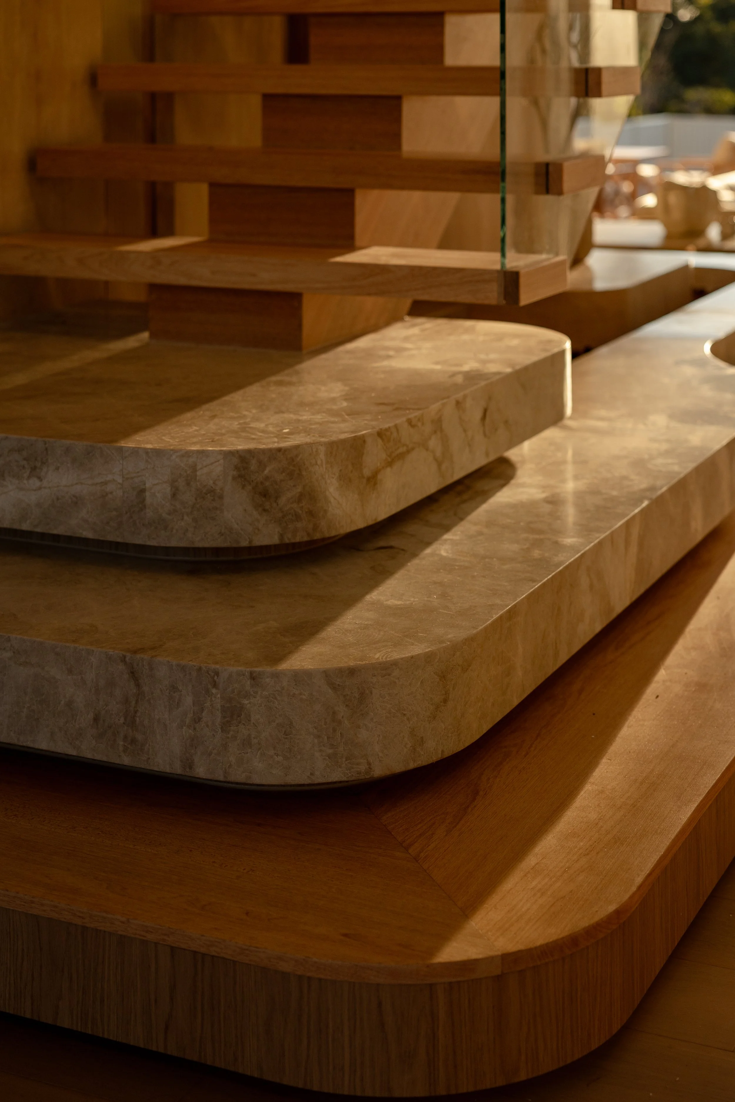 Close-up of a modern wooden staircase with a glass railing, wooden steps, and a curved marble base, with outdoor sunlight and blurred background.
