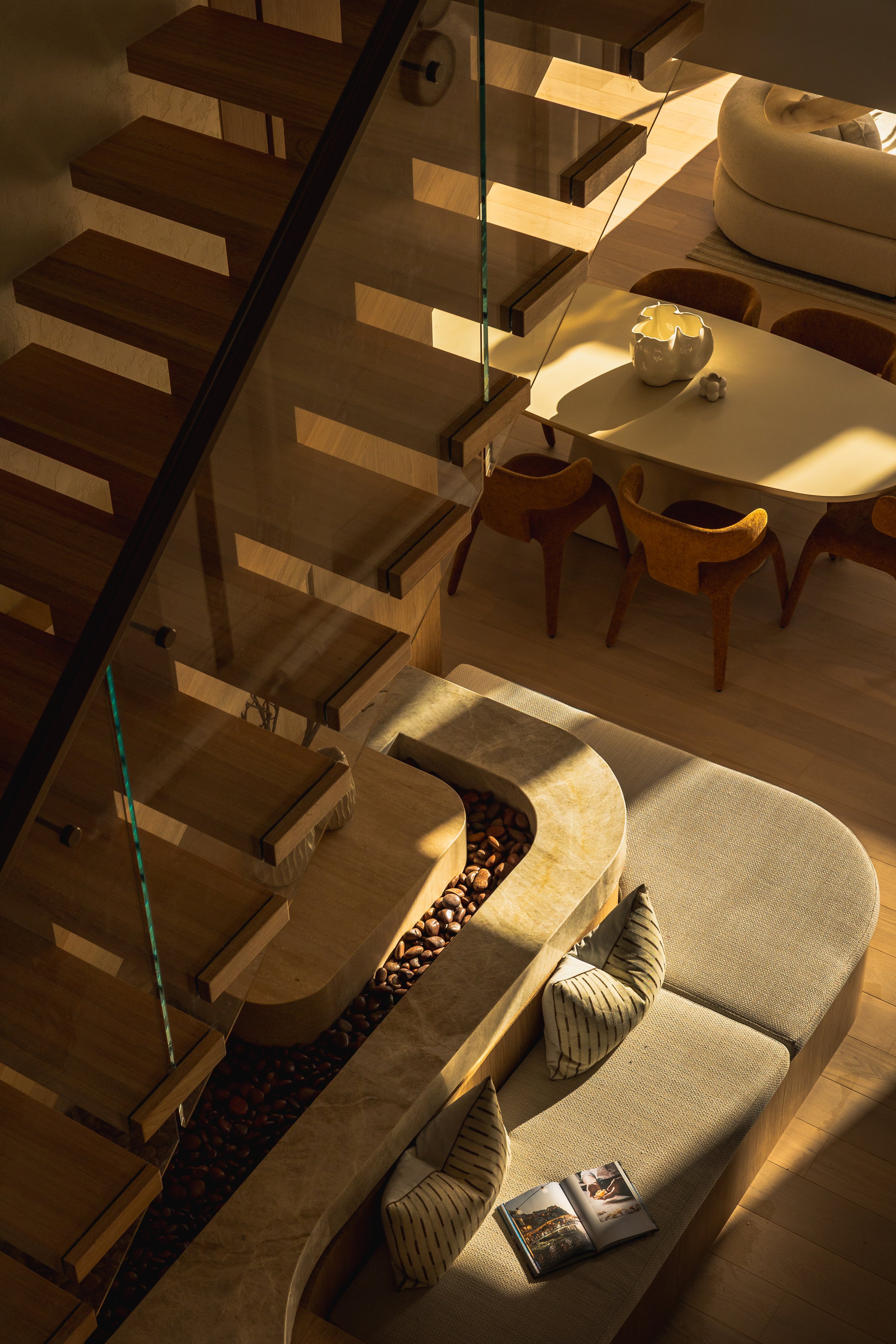 An overhead view of a modern, open-concept living and dining area featuring a staircase with wooden steps and glass railings, a dining table with chairs, a sofa, a decorative vase, and a magazine on a cushioned bench, all illuminated by warm lighting