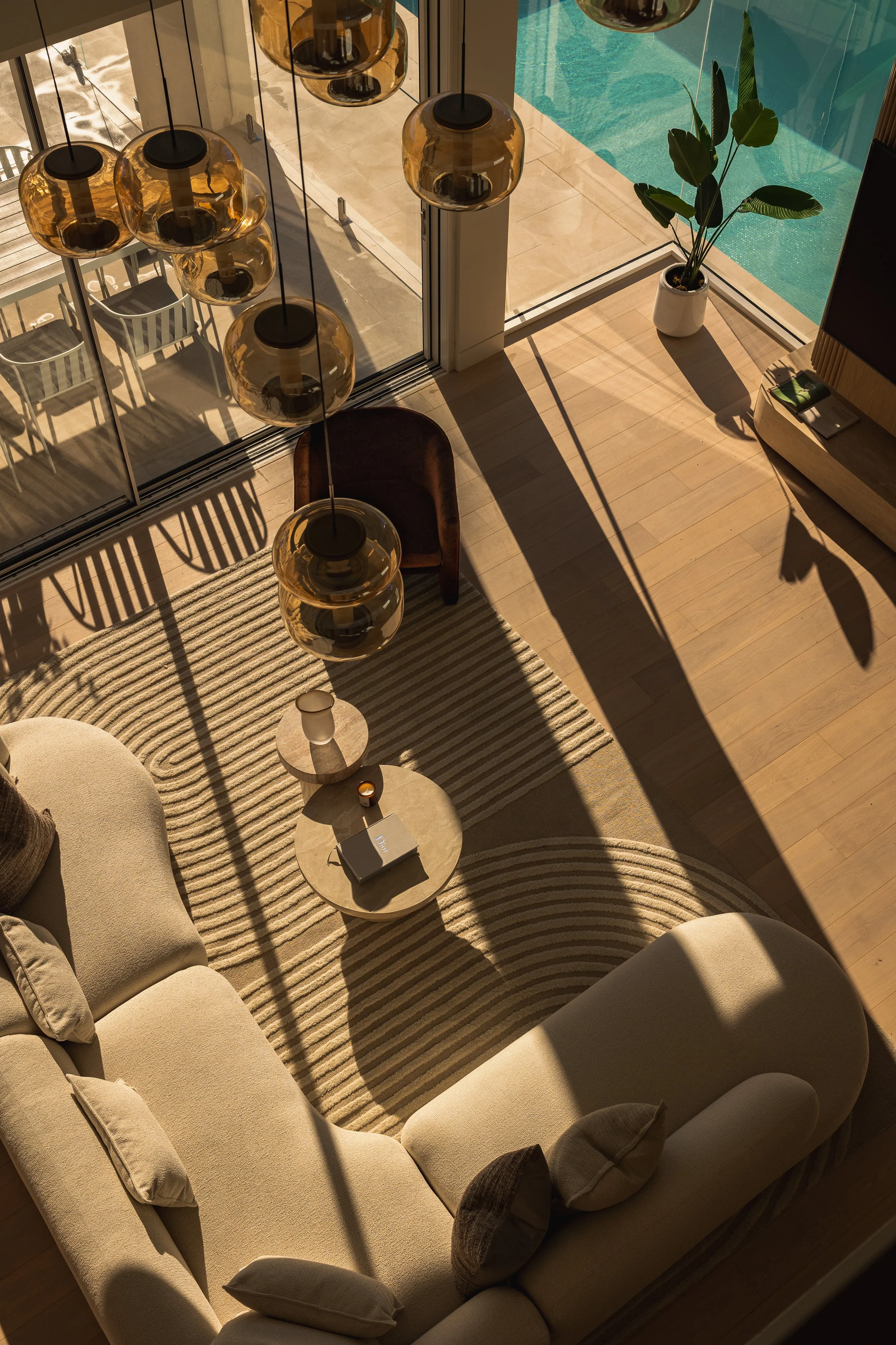 Living room with beige sofa, round coffee table with notebook and decorative item, striped rug, hanging pendant lights with amber glass, sliding glass door opening to outdoor patio with chairs and pool area, potted plant, sunlight creating shadows.