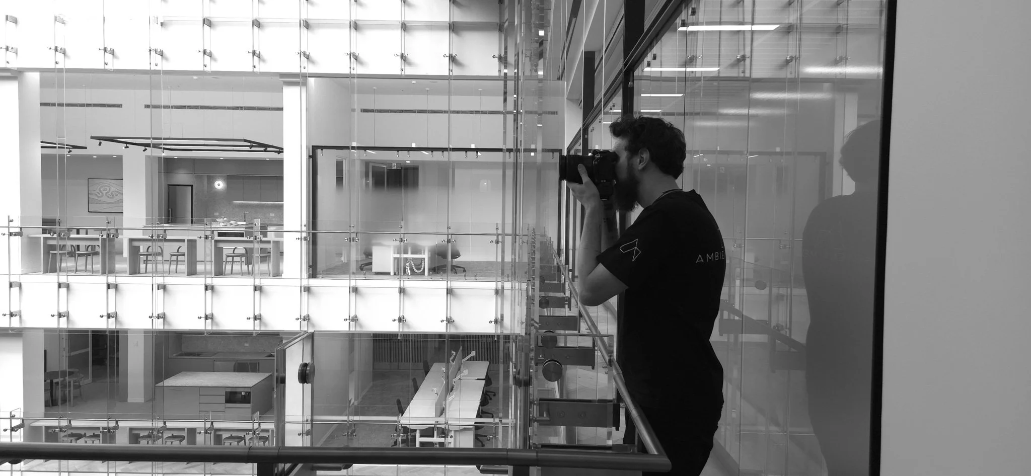 A man with a beard and curly hair holding a professional camera, taking a photograph in a modern, glass-walled office building. The office has multiple levels with desks, chairs, and artwork visible through the glass. High end property development