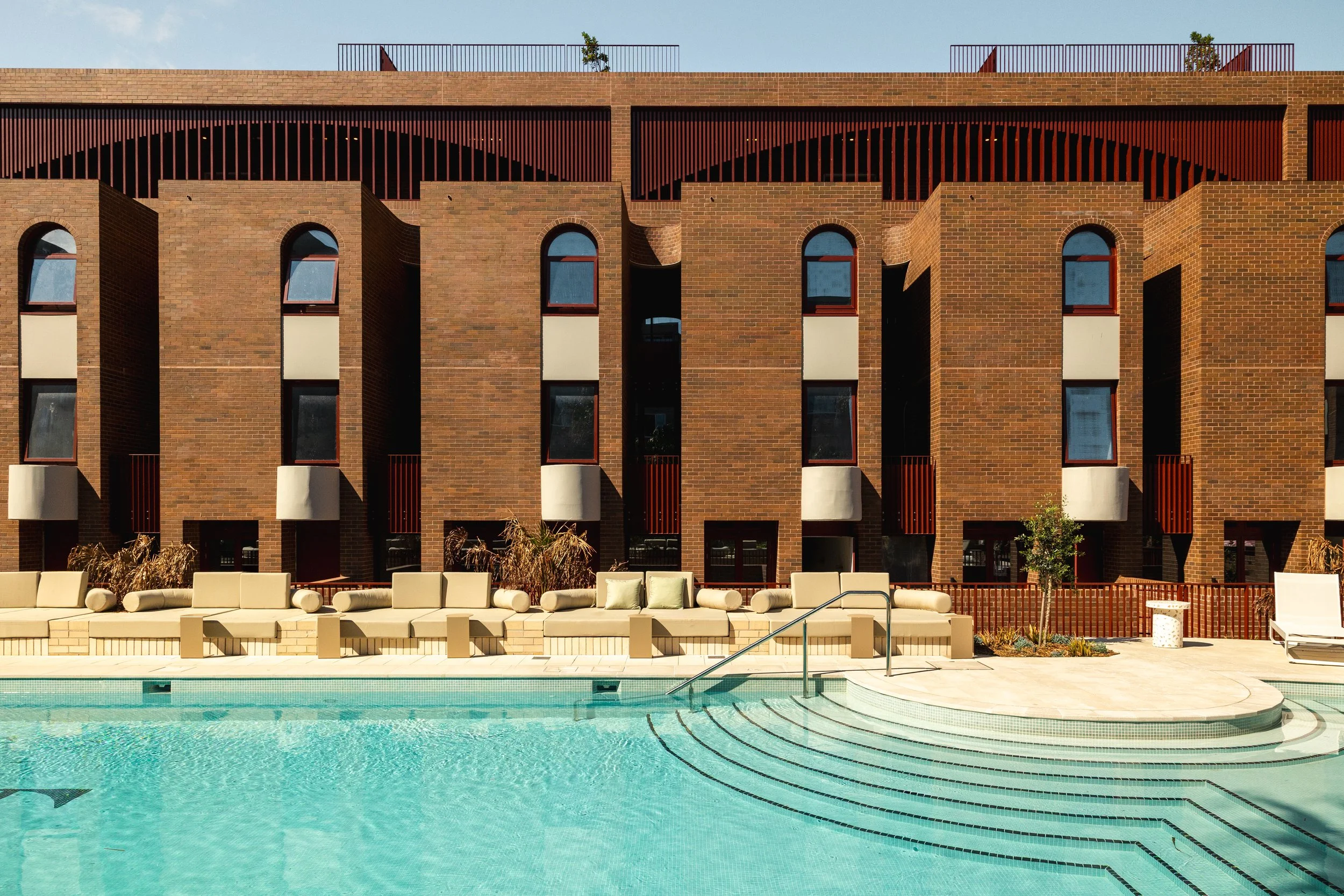 A swimming pool with light blue water in front of a modern brick building with multiple windows and balconies. There is outdoor seating and some plants around the pool.  Luxury architecture design, high end residential development