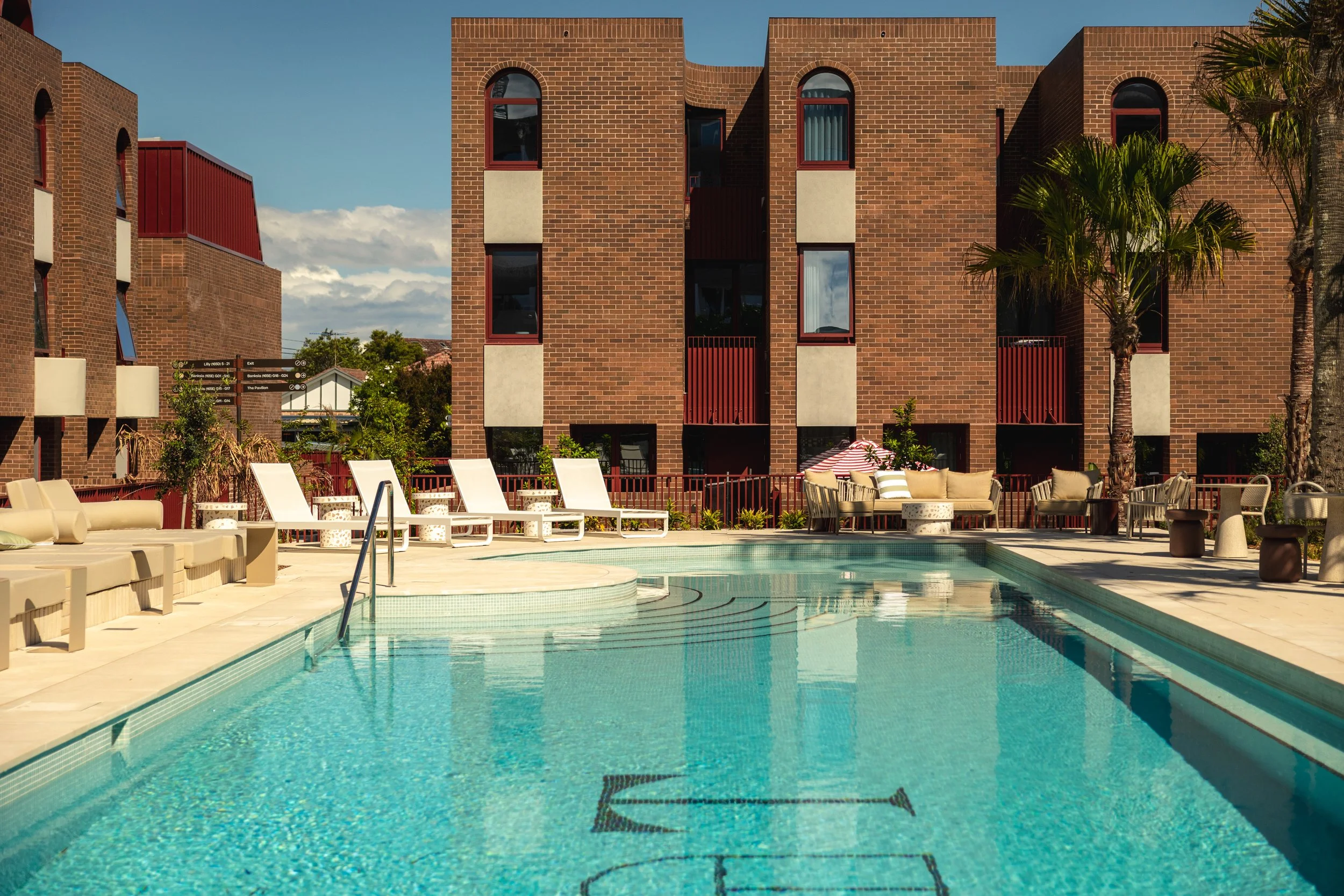 An outdoor swimming pool with lounge chairs and patio furniture, surrounded by brick apartment buildings and palm trees under a blue sky.  Luxury architecture design, high end residential development