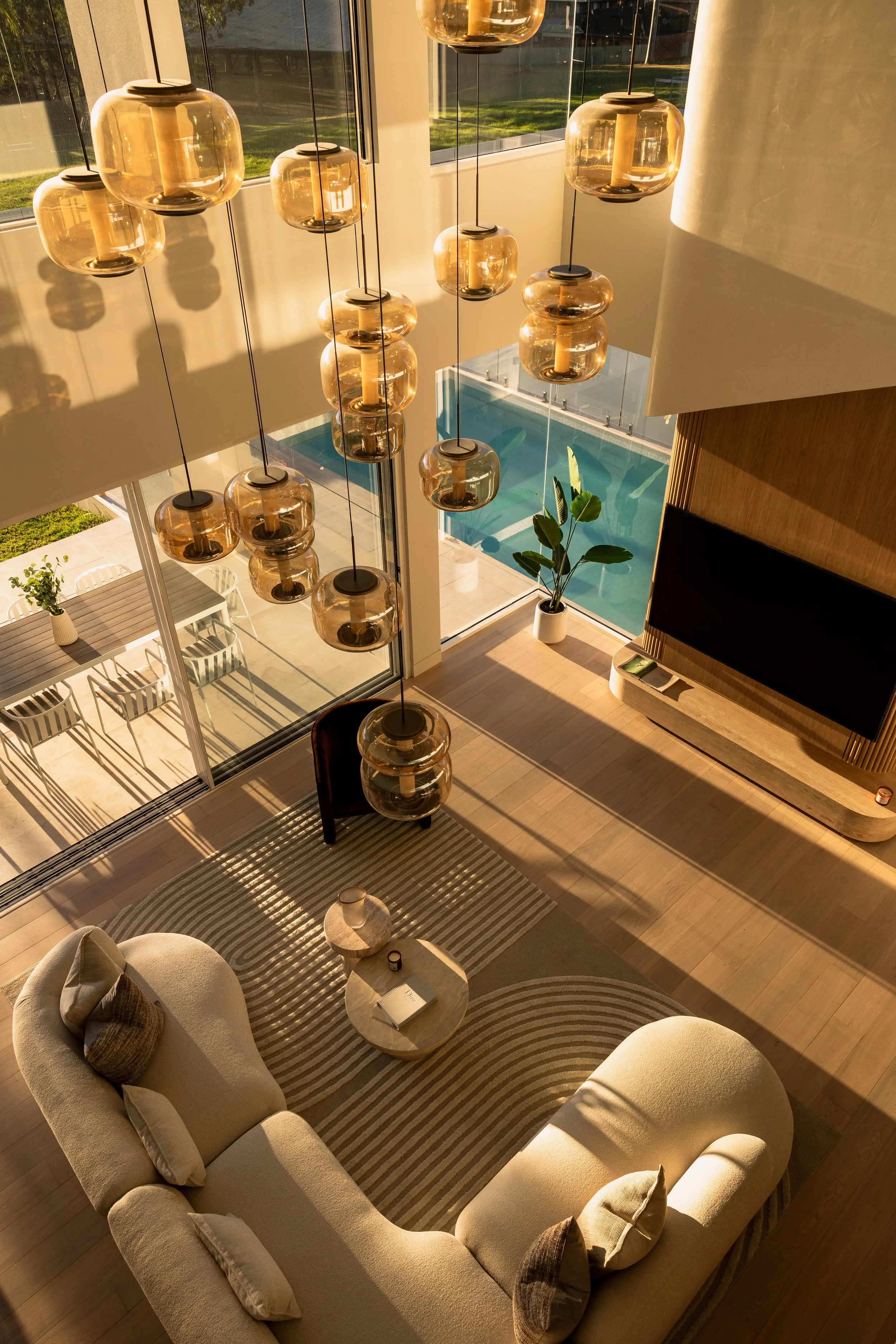 Living room with beige sofa, coffee table, and a striped rug. Large windows with a view of a pool and outdoor patio. Hanging pendant lights with glass covers illuminate the room.