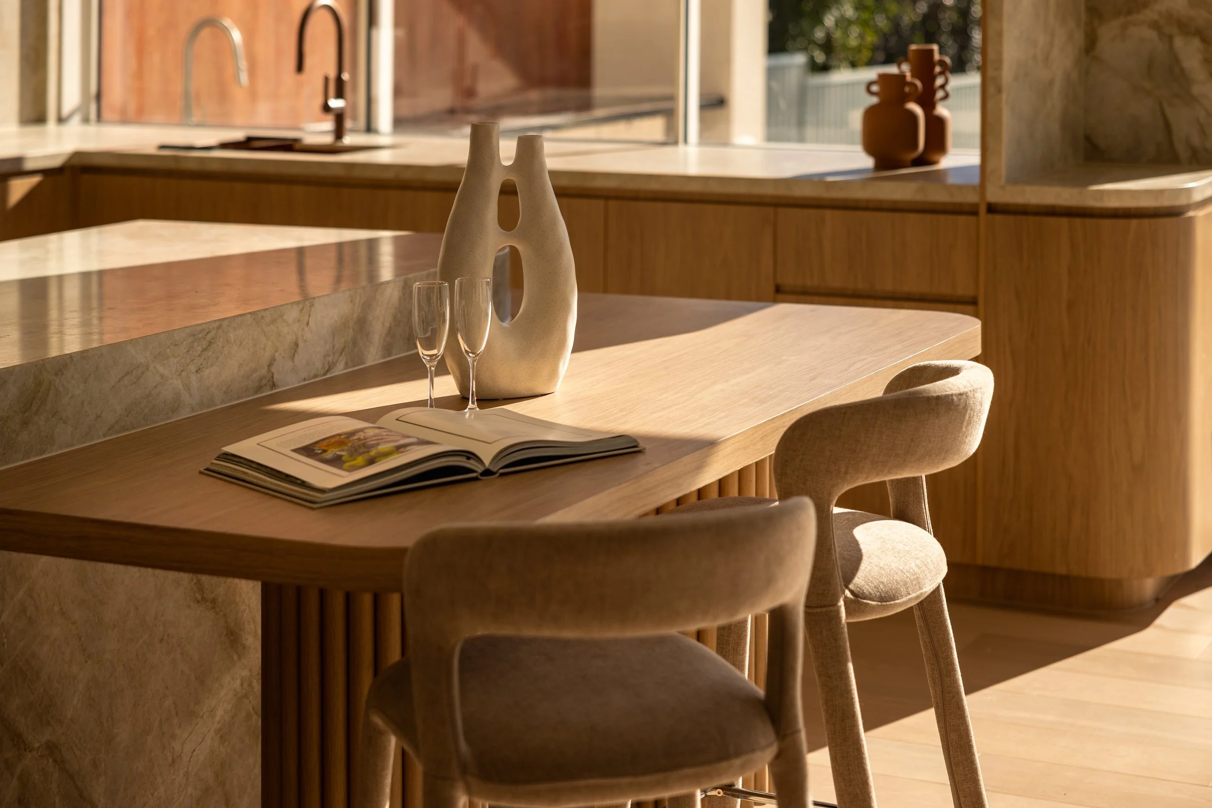 A modern kitchen with a wooden dining table, beige upholstered chairs, a decorative vase with a cutout design, two empty wine glasses, and an open magazine on the table. Sunlight streams in through a window.
