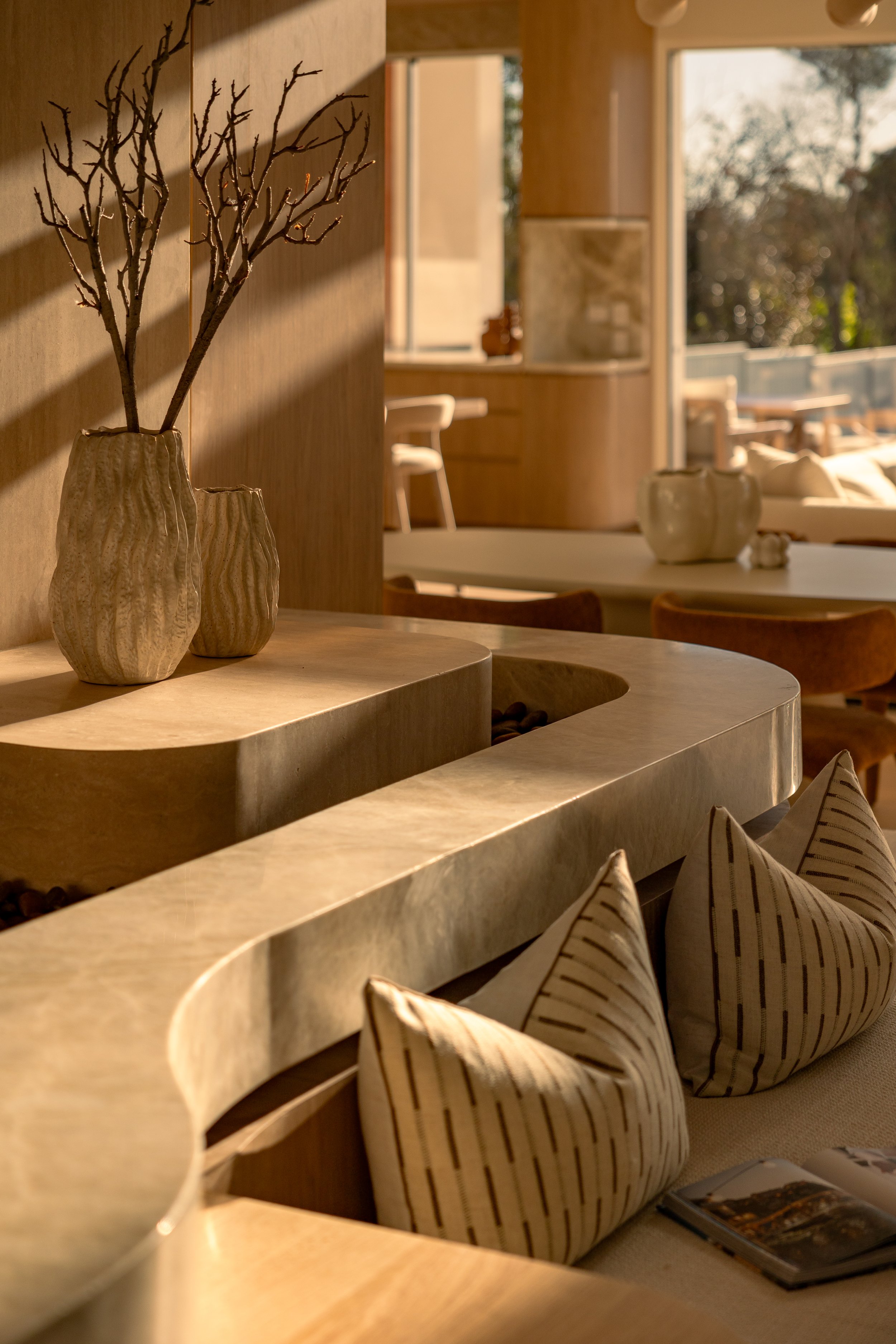 Interior of a room with beige and wooden furniture, decorative vases on a curved wooden countertop, patterned coffee table books, and pillows with pinstripe design.