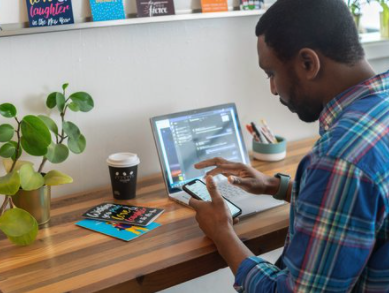 Man wearing a plaid shirt using a smartphone at a wooden desk, with a laptop, a coffee cup, and a magazine, in a room with a white wall and potted plant.