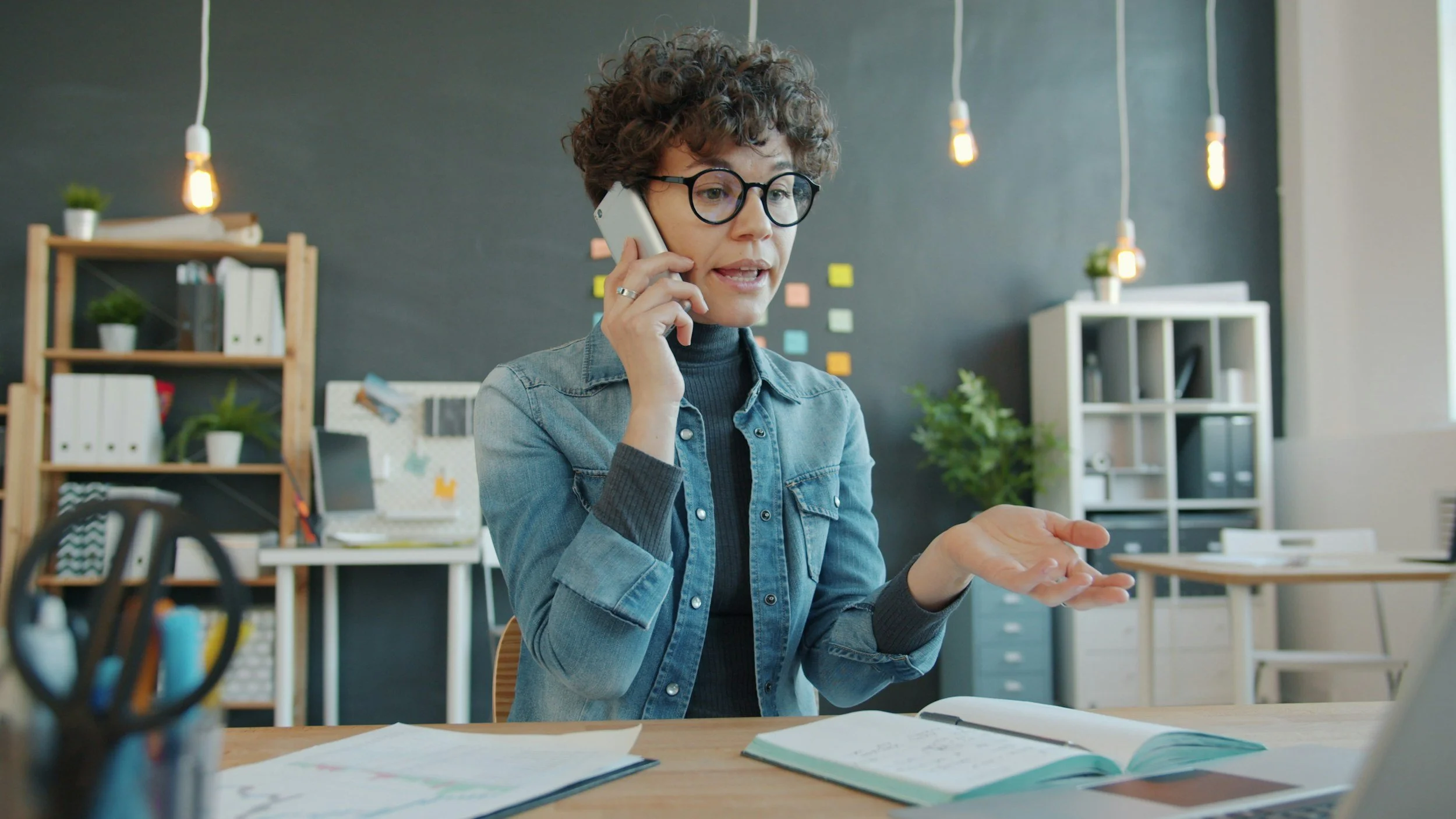 Woman with curly hair and glasses talking on a cellphone at a desk in an office, gesturing with her hand, with office supplies and shelves in the background.