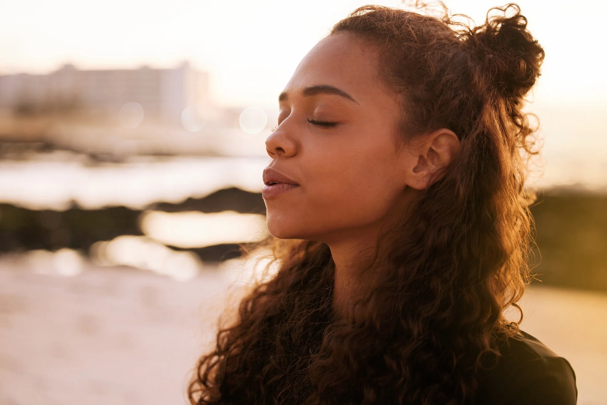 A woman with curly red hair, eyes closed, faces a sunset by the water.