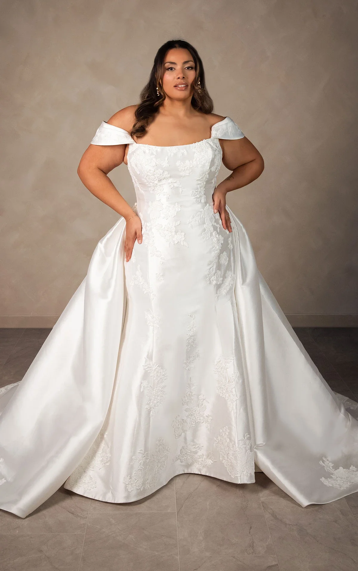 A woman in a white wedding gown with off-shoulder sleeves, floral embroidery, and a full train, standing against a neutral background.