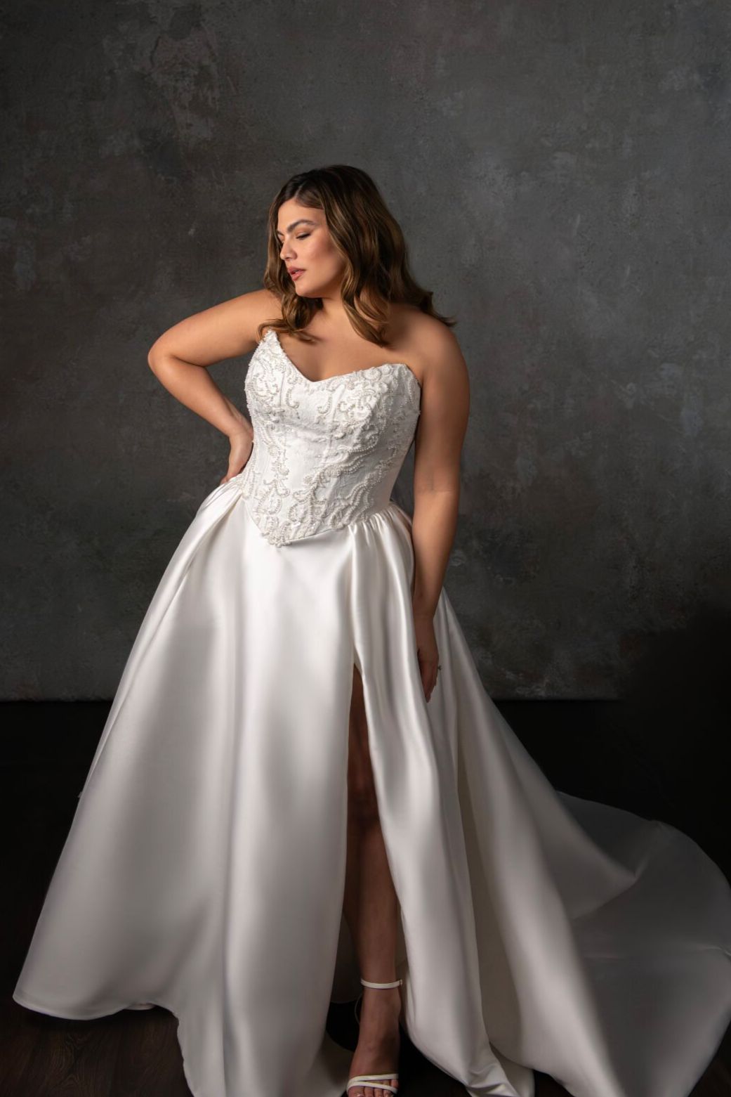 A woman wearing a white bridal gown with lace details on the bodice, standing against a dark textured wall. The dress has a high slit, and she is wearing high-heeled sandals. She has brown, shoulder-length, wavy hair, and is posing with one hand on her hip and her eyes closed.