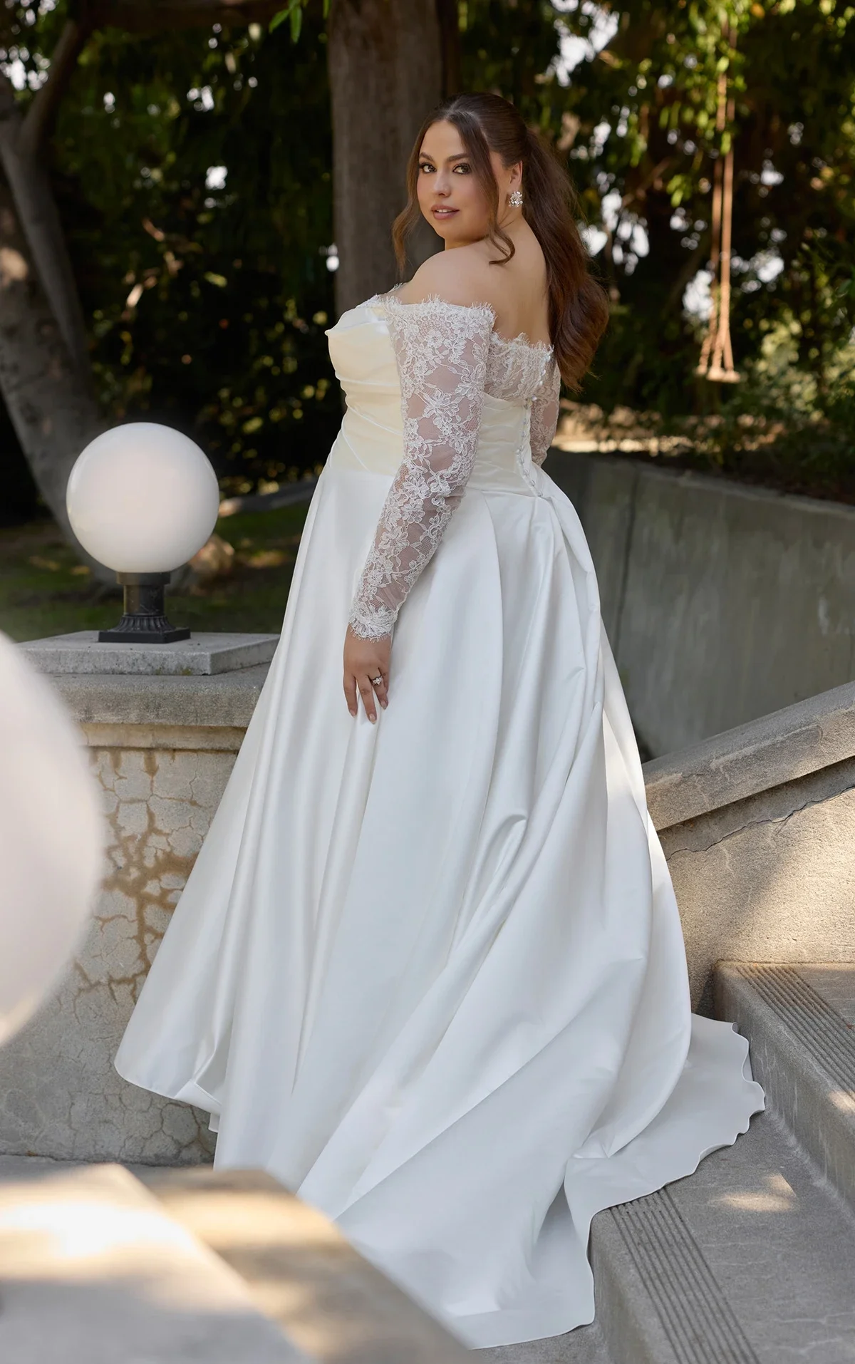 A woman in a white wedding gown with lace long sleeves and off-the-shoulder neckline, standing outdoors on a staircase with greenery in the background.