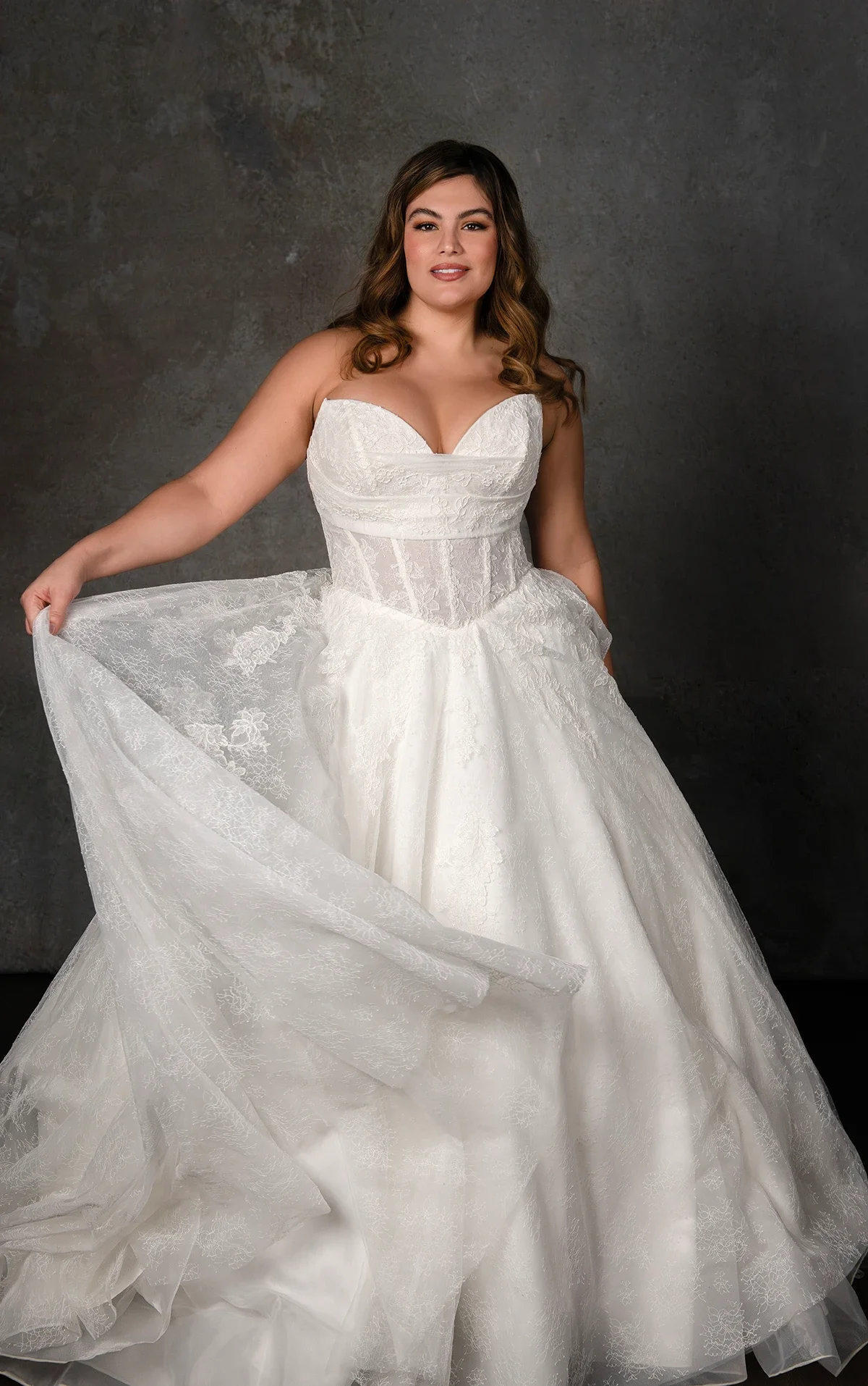 A woman in a strapless white wedding gown with lace details, standing against a dark textured background, holding the skirt of her dress.
