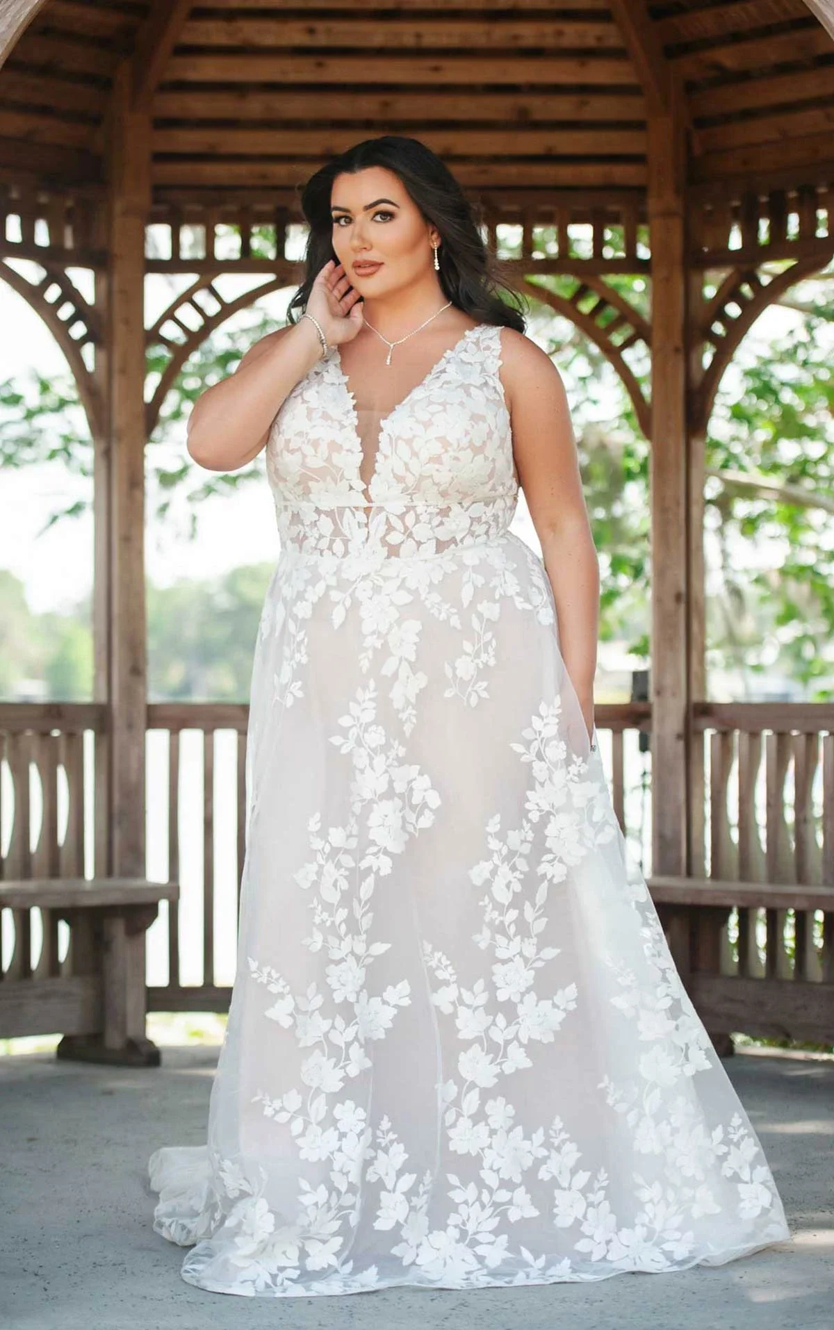 A woman in a white lace wedding dress with floral embroidery, standing in a wooden gazebo with trees in the background.