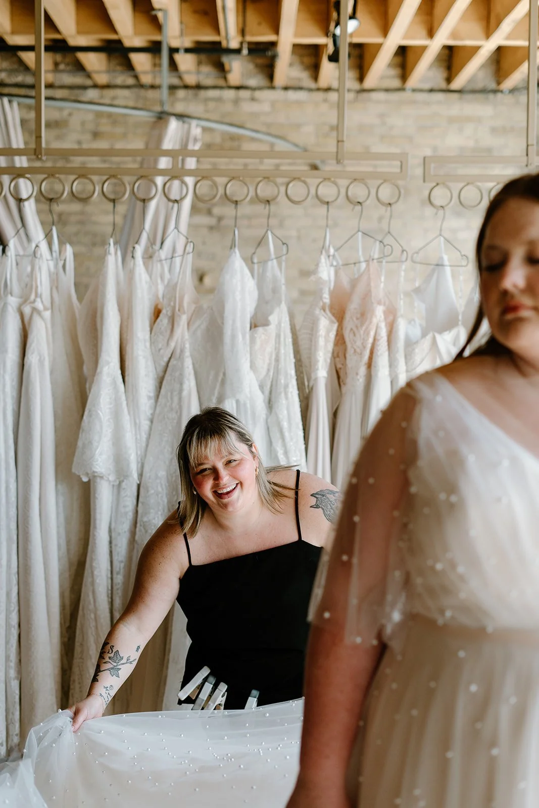 Two women in a bridal boutique with wedding dresses hanging on racks behind them. The woman on the left, with a smile, is wearing a black top and helping the woman on the right, who is wearing a wedding dress with pearl embellishments.
