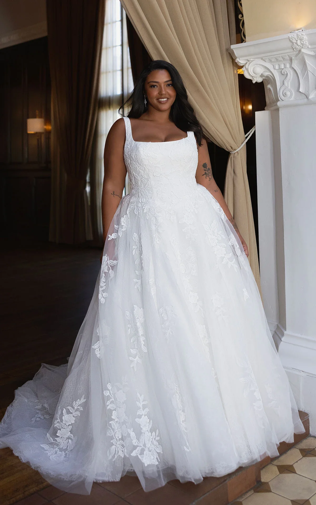A woman in a white wedding dress with lace details, standing indoors near a decorative fireplace with beige curtains in the background.