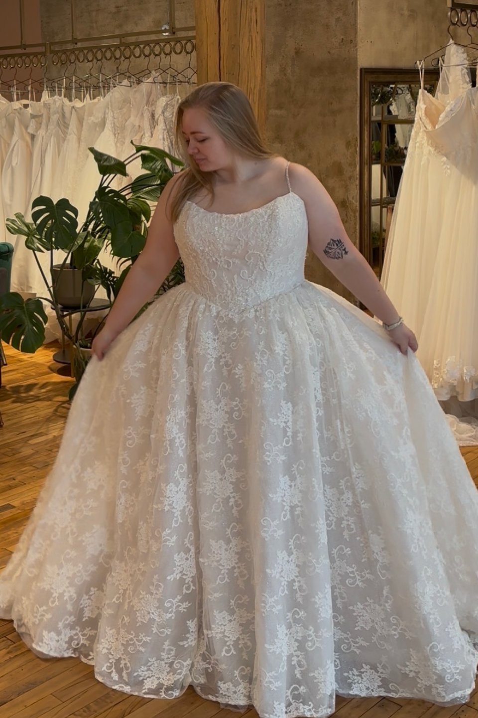 A woman in a white, lace wedding dress trying it on in a bridal store, holding out the sides of her dress. The store has a wooden floor, a mirror, and several other wedding dresses hanging in the background.
