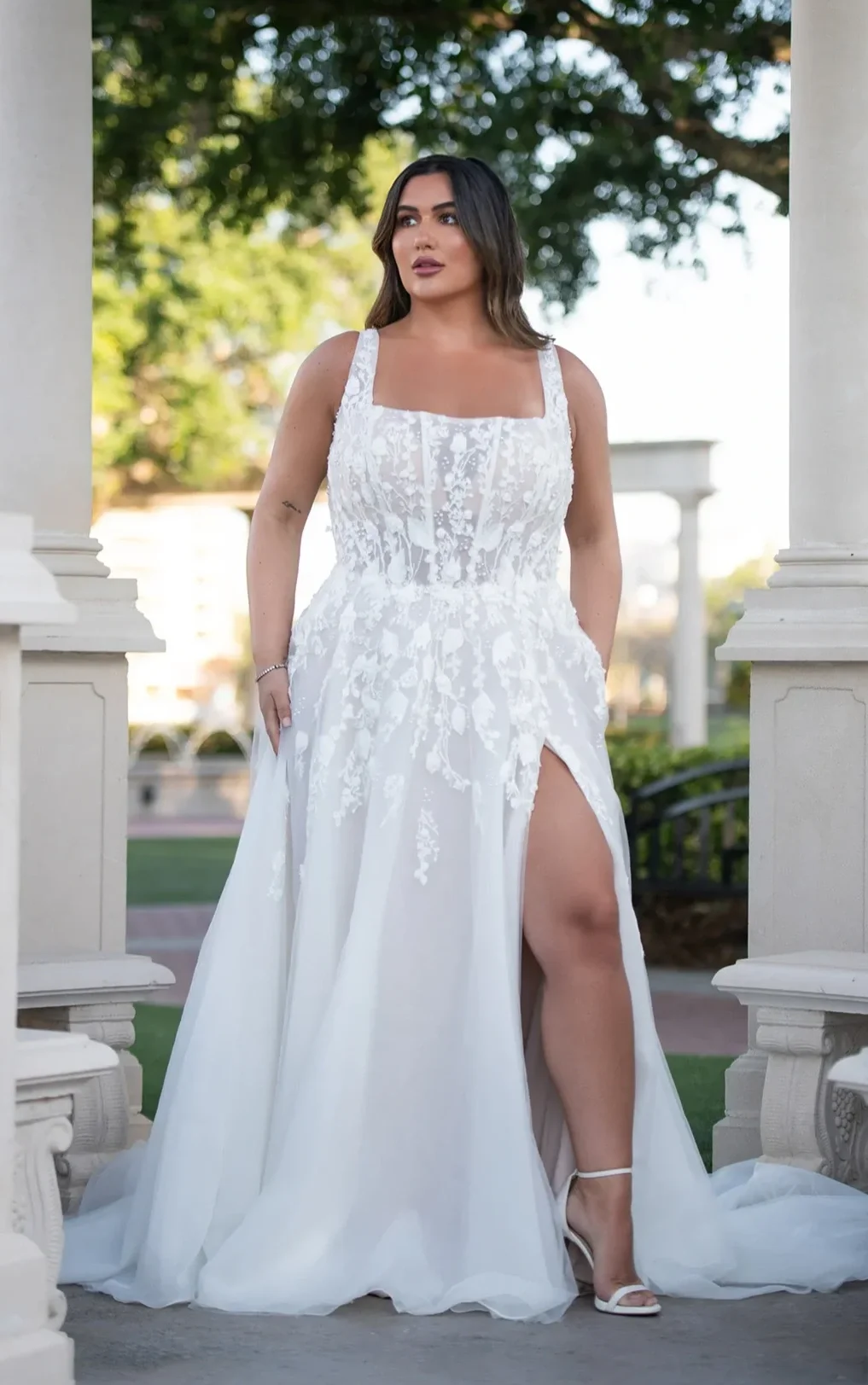 A woman in a white wedding dress with floral lace details, standing outdoors between white pillars with a background of trees and a white structure, posing confidently with one leg slightly raised, revealing her white high-heeled sandals.