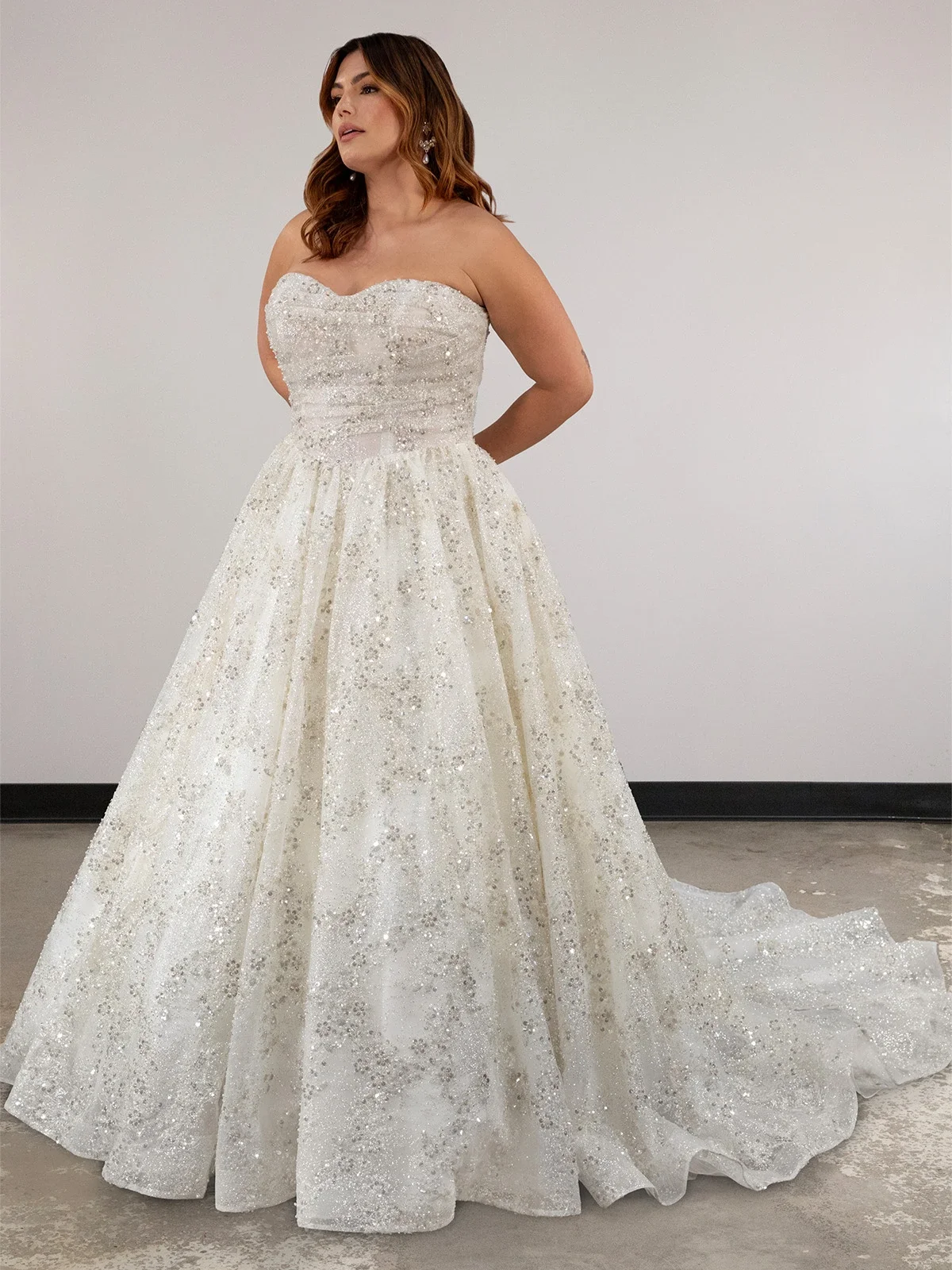 A woman in a strapless, sparkling white wedding gown with a full-length skirt and train, standing against a plain white wall.