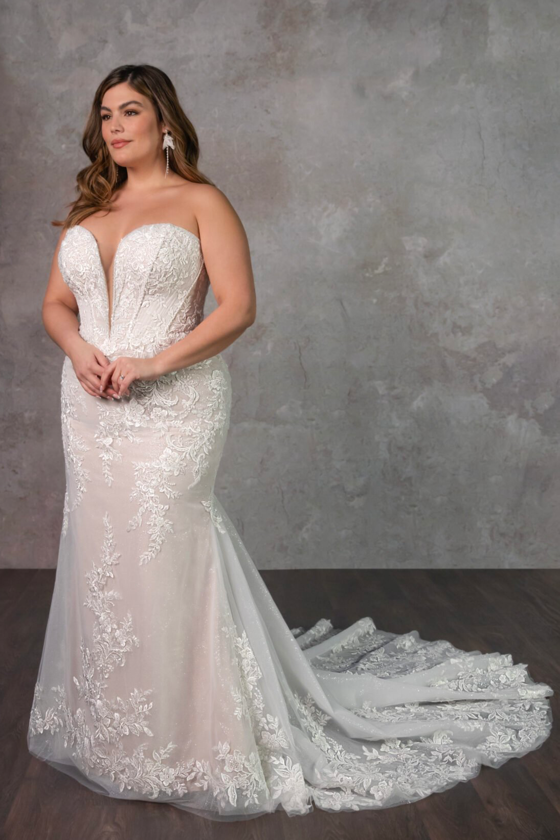 A woman wearing a lace wedding gown with a strapless sweetheart neckline and a train, standing against a textured grey wall.