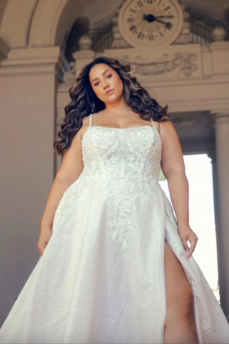 Woman in a white wedding dress with spaghetti straps, standing in a grand interior with a clock on the wall behind her