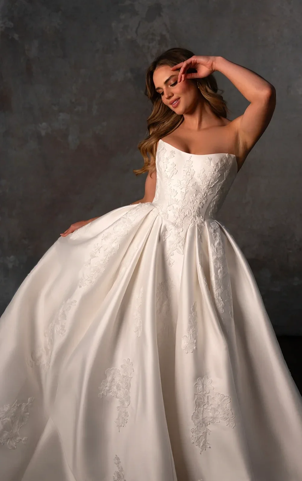 A woman in a strapless white wedding gown with floral embroidery, standing against a grey textured background, gracefully posing with her eyes closed and hand near her face.