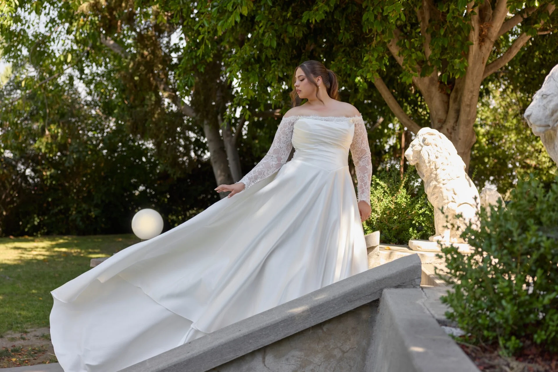 far away photo of plus size bride wearing ivory satin aline wedding dress with basque waist and lace off the shoulder long sleeves showing off long train