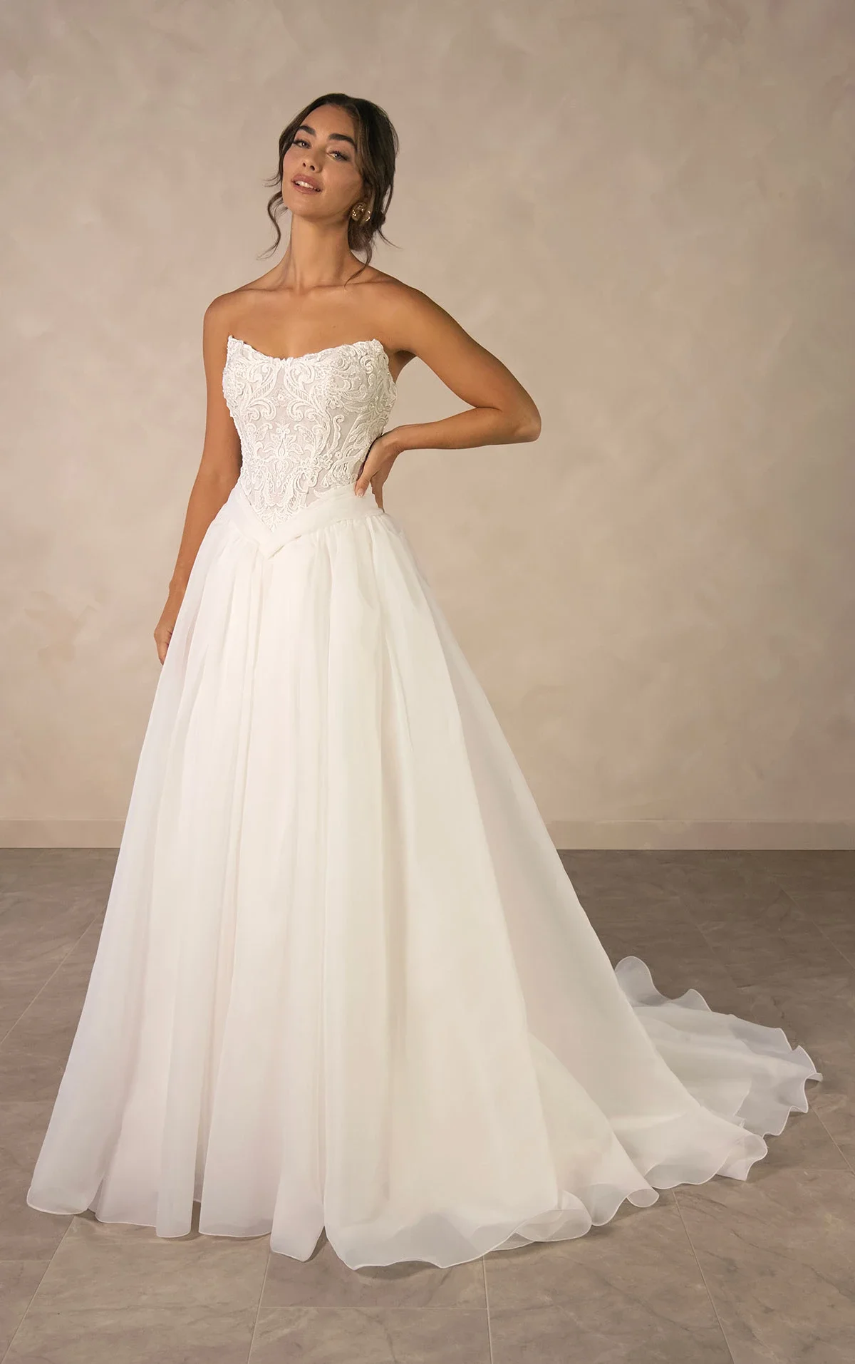 A woman wearing a strapless white wedding gown with lace bodice and full flowing skirt, standing on a beige tiled floor against a plain beige wall.