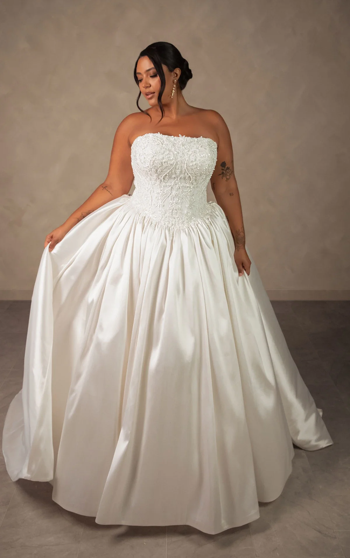 Woman in a strapless wedding gown with lace and embroidery details on the bodice, holding the skirt, standing against a neutral background.