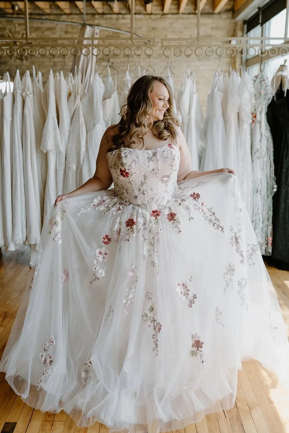Woman trying on a wedding dress in a boutique, surrounded by other white wedding gowns hanging on a rack.