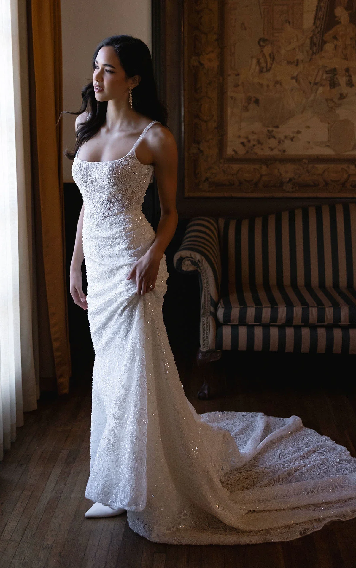 A woman in a white, lace wedding dress with beaded details stands indoors near a window, looking out. She has dark, wavy hair and is wearing long, dangling earrings, with a decorative rug hanging on the wall behind her and a striped armchair nearby.