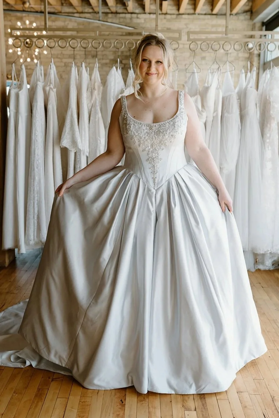 A woman trying on a wedding dress in a bridal boutique, surrounded by other wedding gowns on hangers. She is holding the sides of her dress and smiling.