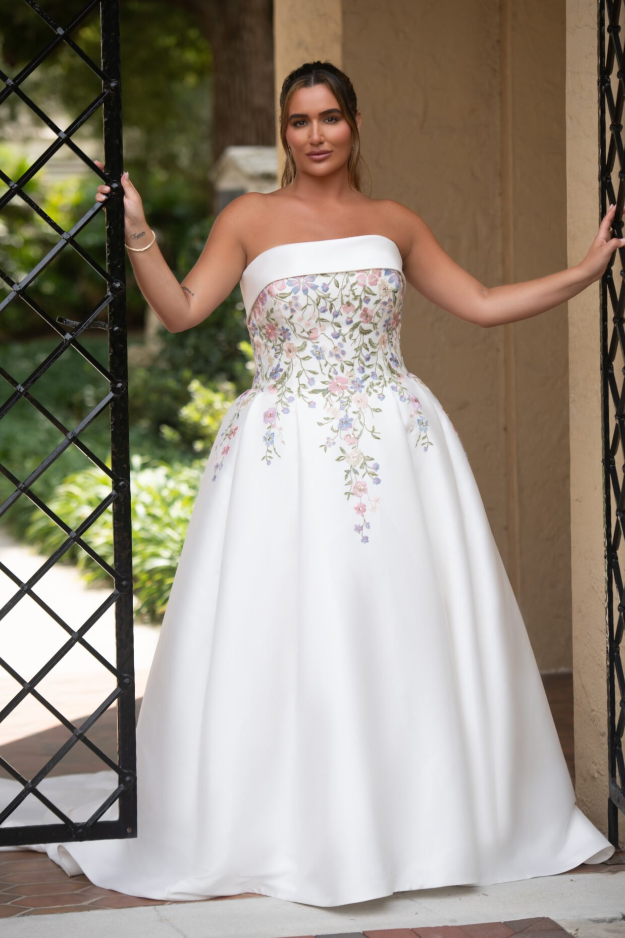Woman in a strapless white floral gown standing between black metal gates outside.